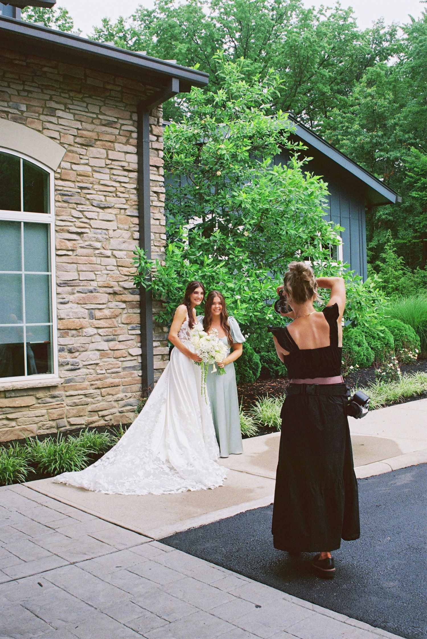 Photographer capturing bride in white gown and bridesmaid in mint dress posing outside stone building surrounded by lush greenery.