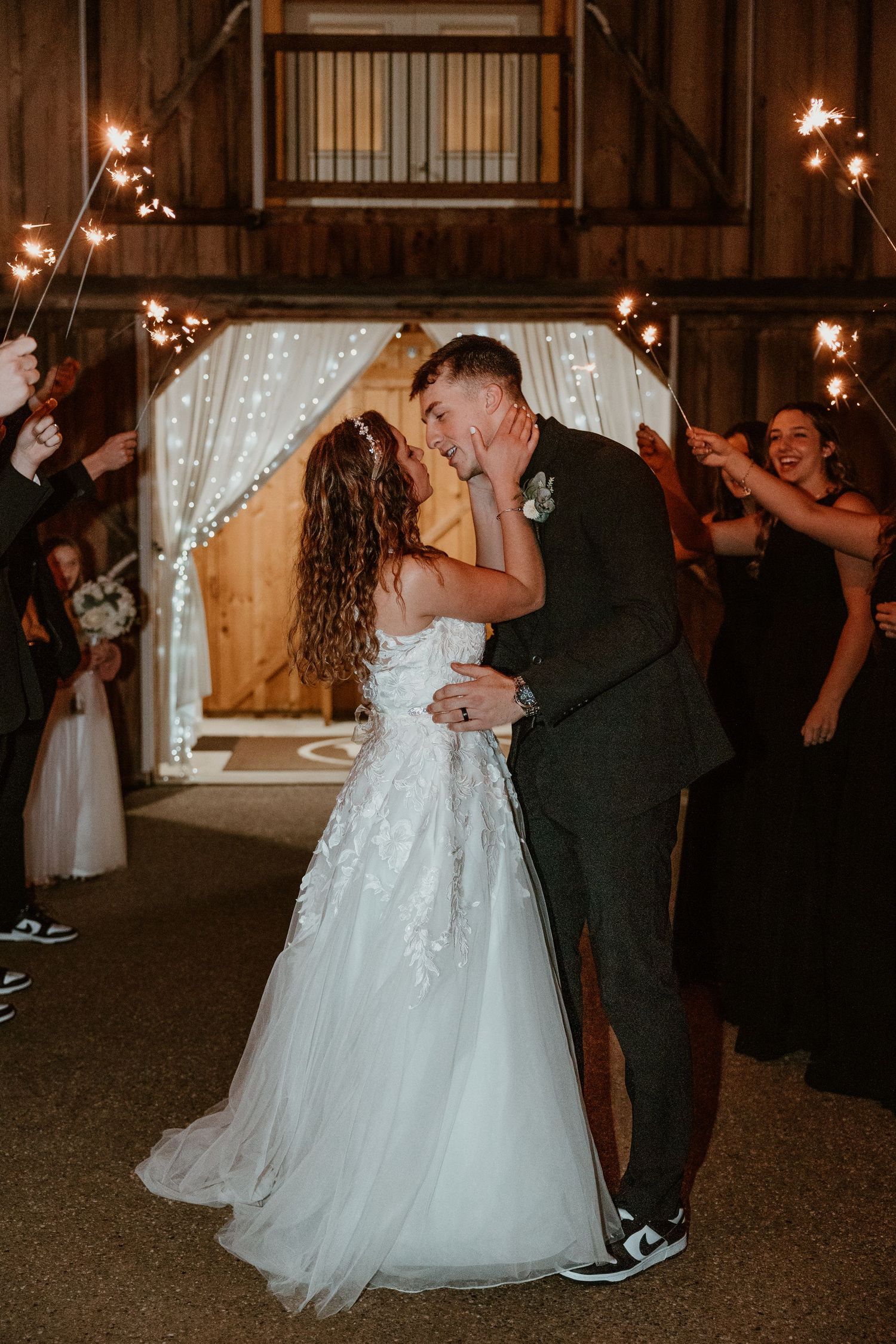 A couple shares a romantic dance in a rustic barn venue with string lights and wooden beams overhead.