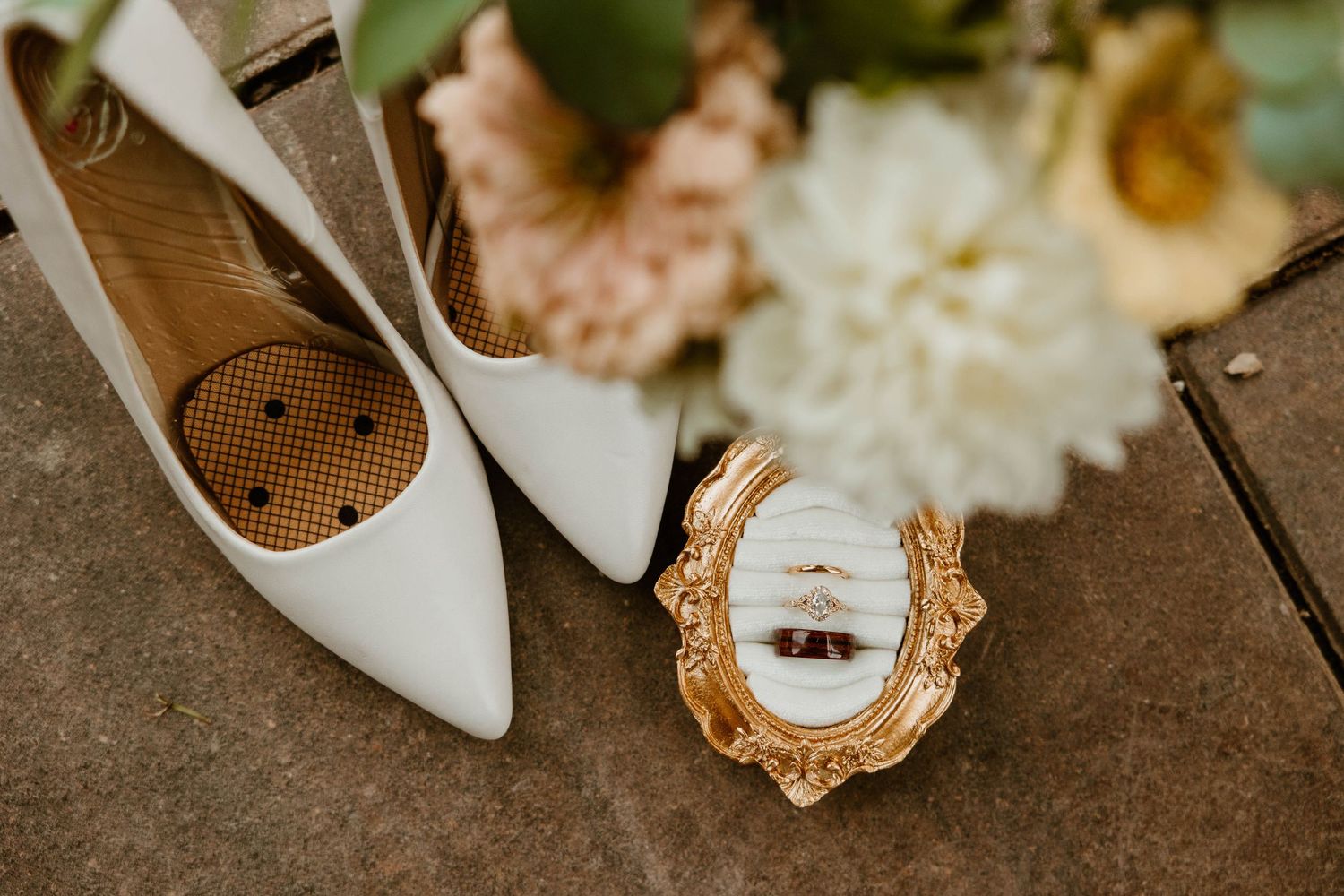 White bridal heels beside a gold ornate ring box with wedding bands and a soft floral bouquet on a rustic surface.