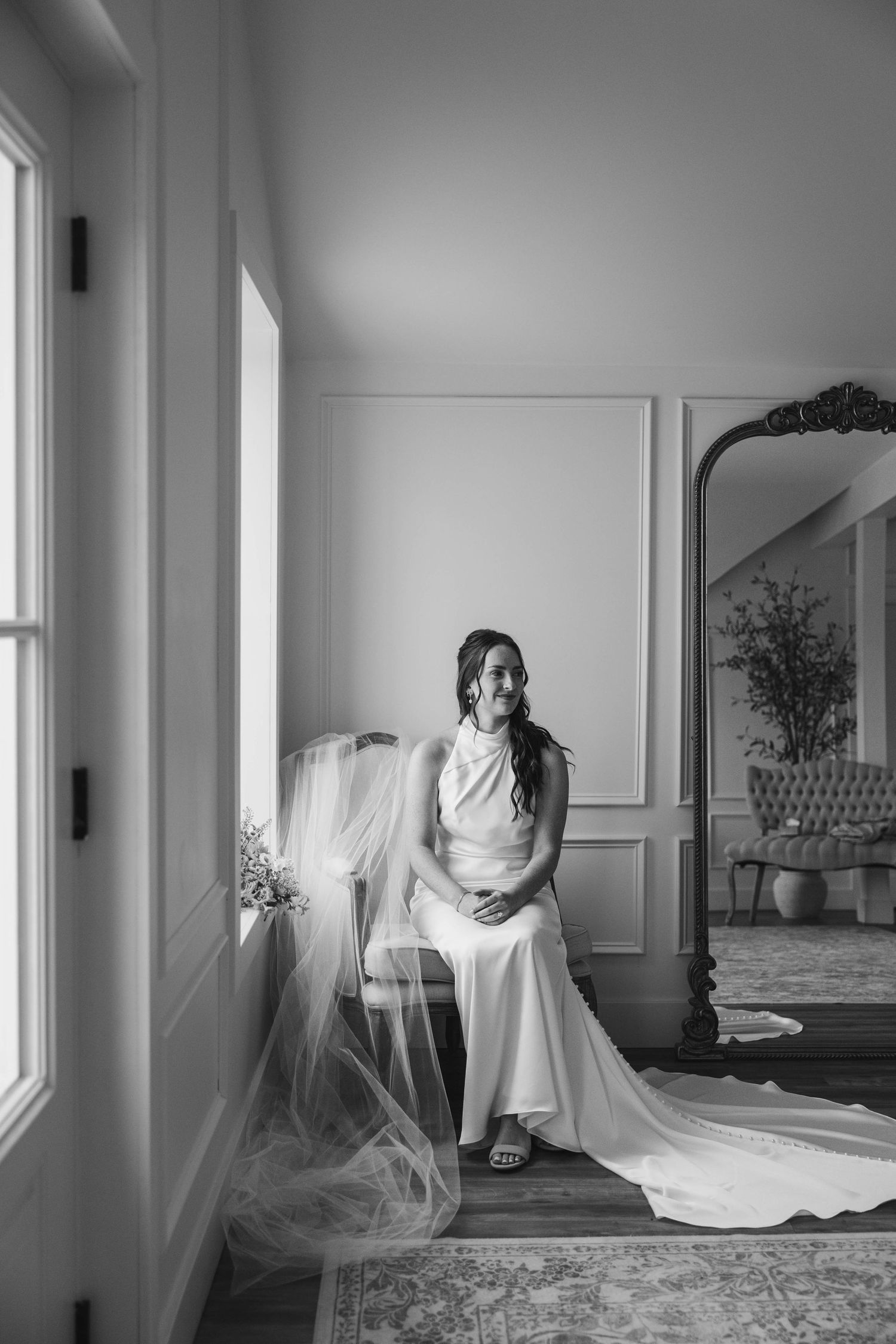 Bride in white gown and veil sits by window in elegant room, black and white wedding portrait.