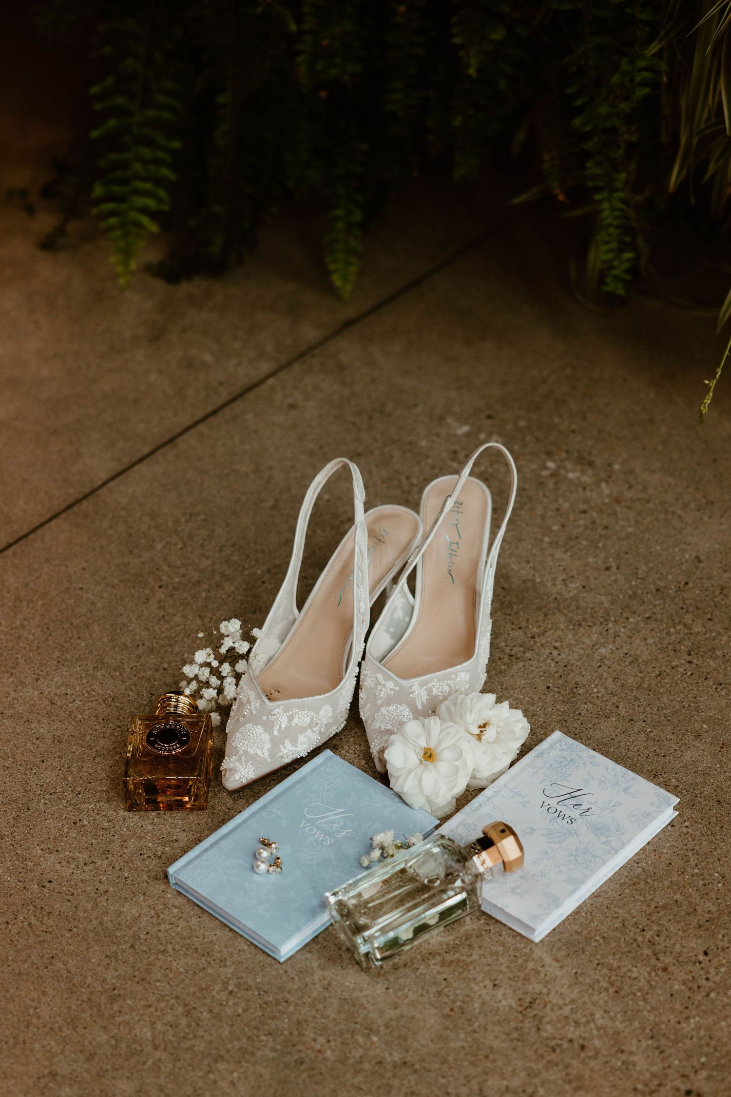 Bridal flat lay featuring white lace heels, perfume, rings, and vow books on a concrete floor.