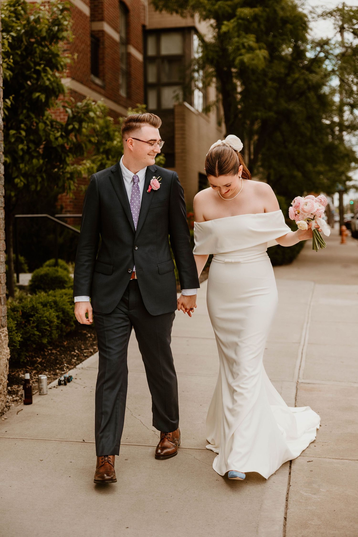 A couple walks and dances together on a sidewalk outside a brick building.