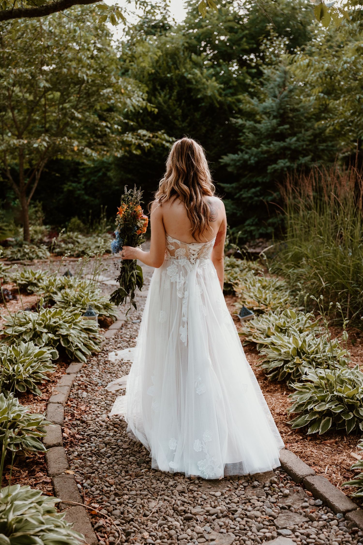 A bride in a flowing white wedding dress walks along a garden path surrounded by lush greenery.