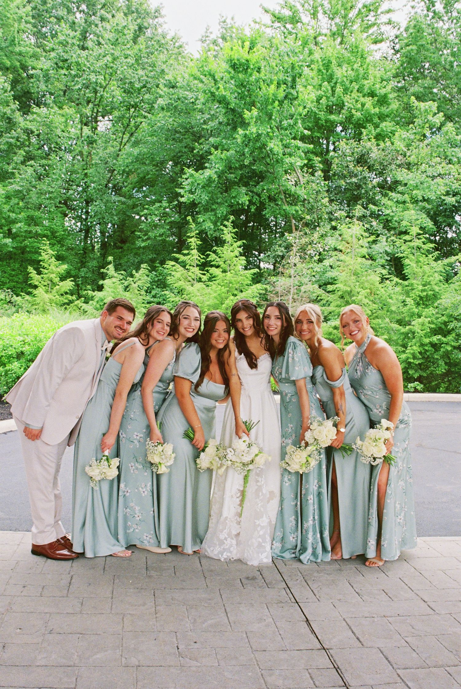 Bride in white lace gown poses with bridesmaids in sage green dresses and groomsman outdoors.