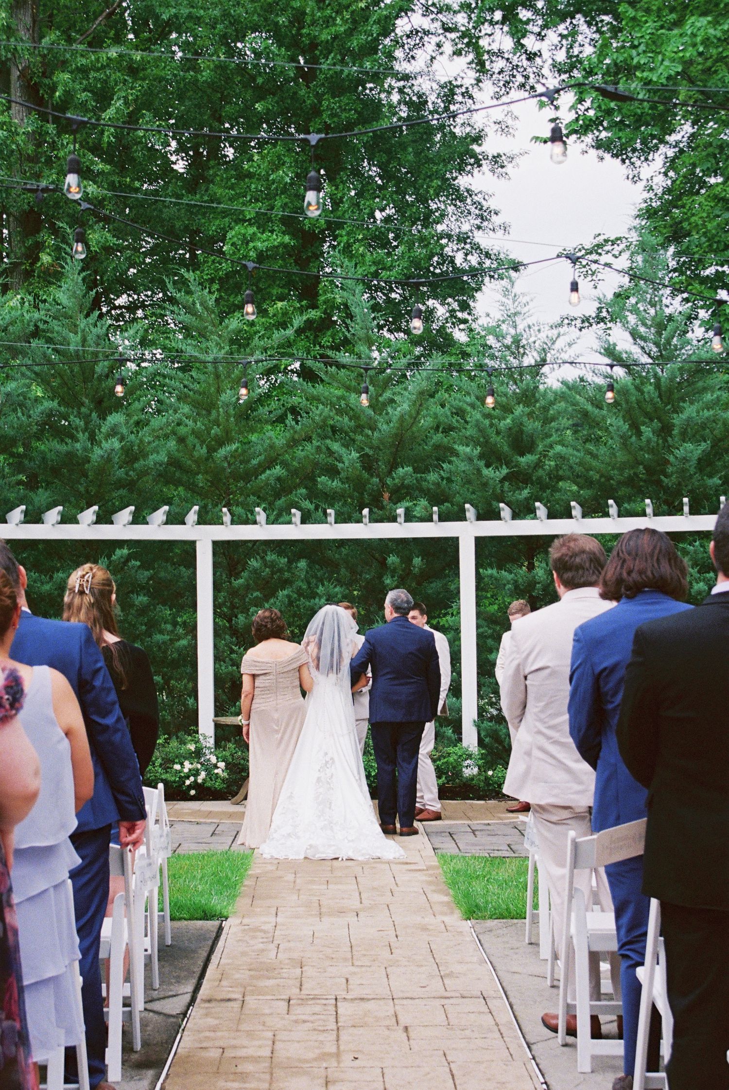 Bride walks down aisle at outdoor garden wedding ceremony surrounded by tall evergreen trees.