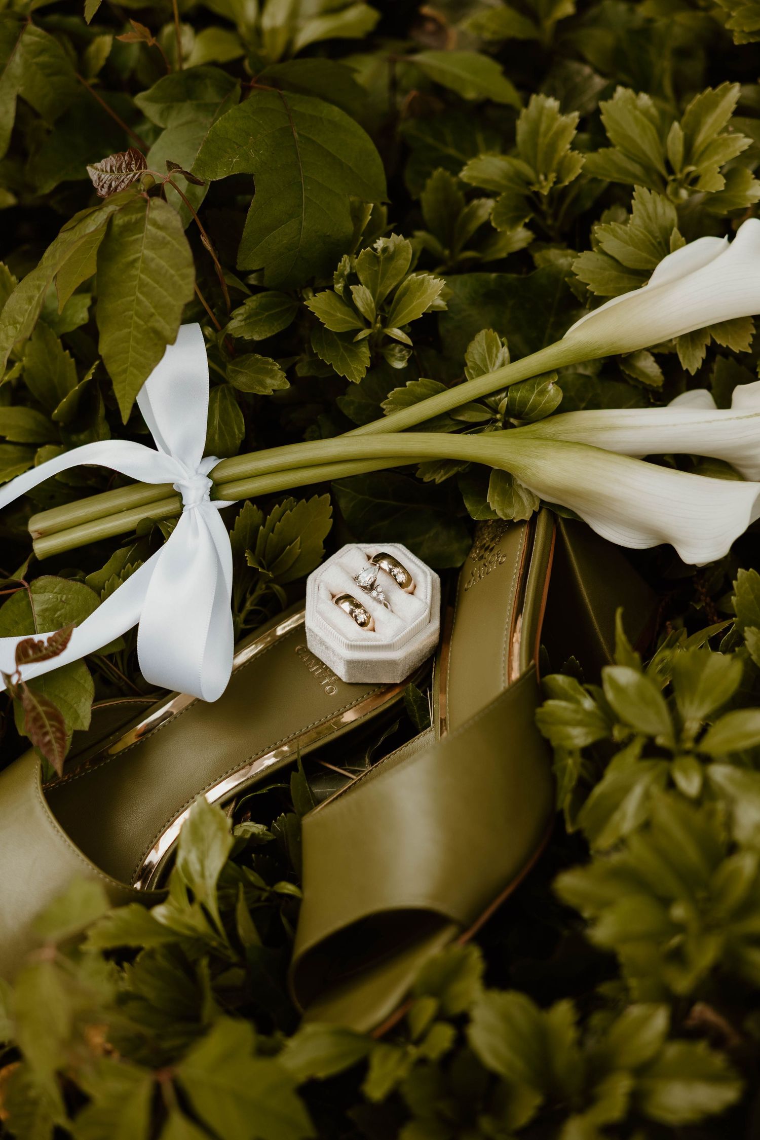 White calla lily bouquet and wedding rings arranged artistically among green foliage.