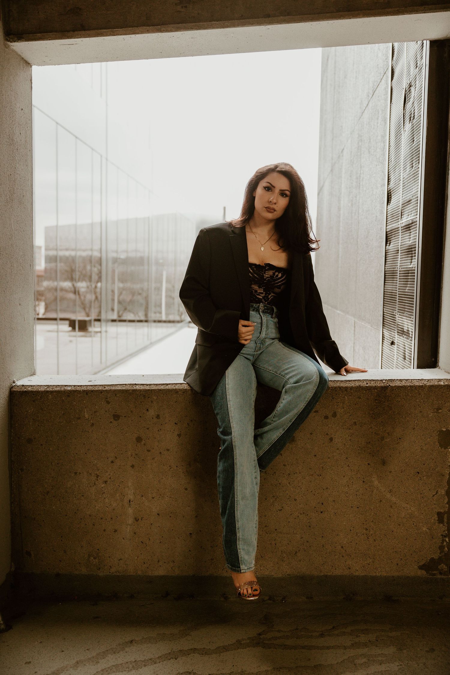 Woman in black jacket poses casually on concrete ledge by glass windows.