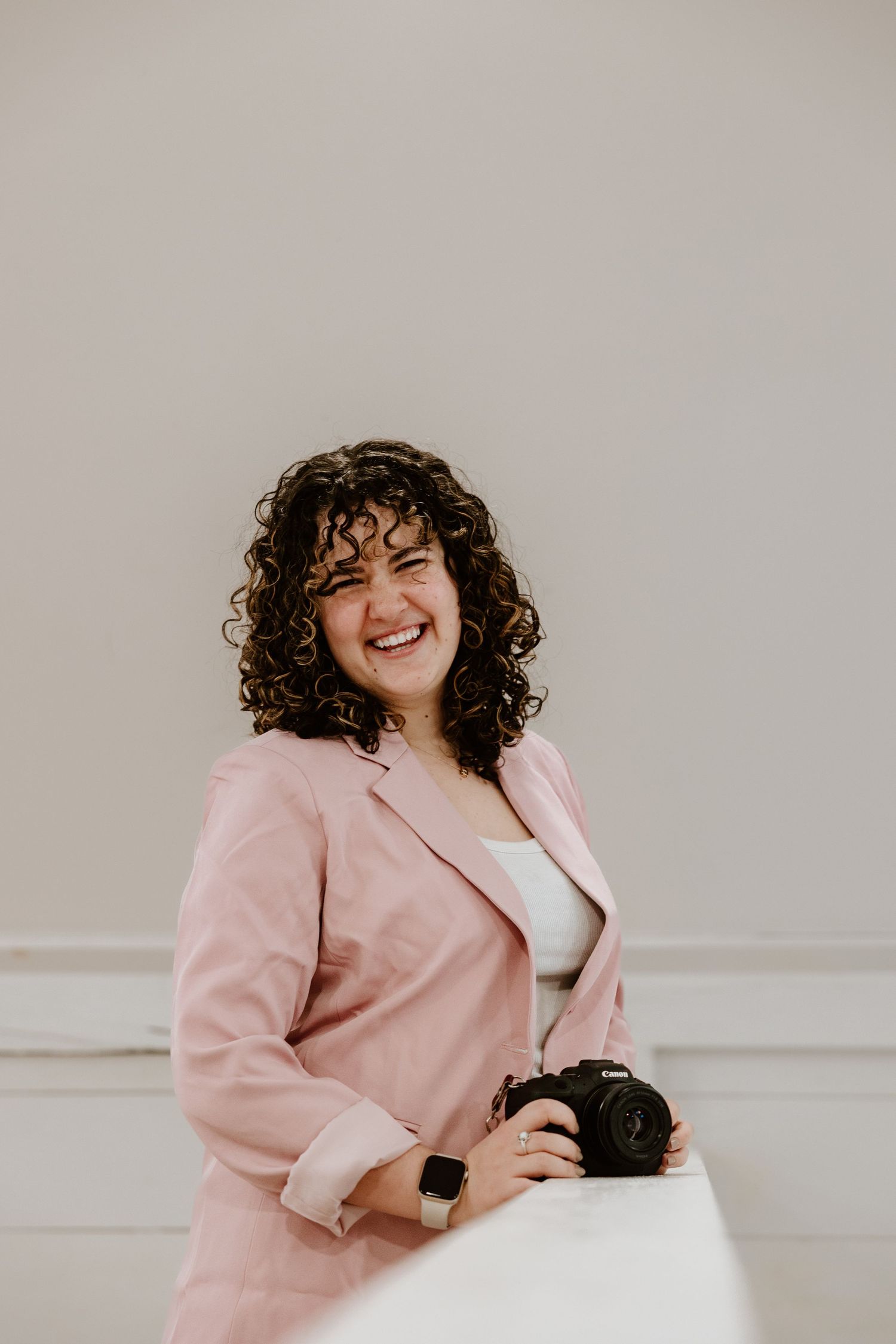 Professional wearing a pink blazer smiles warmly while holding a camera in a light studio setting.