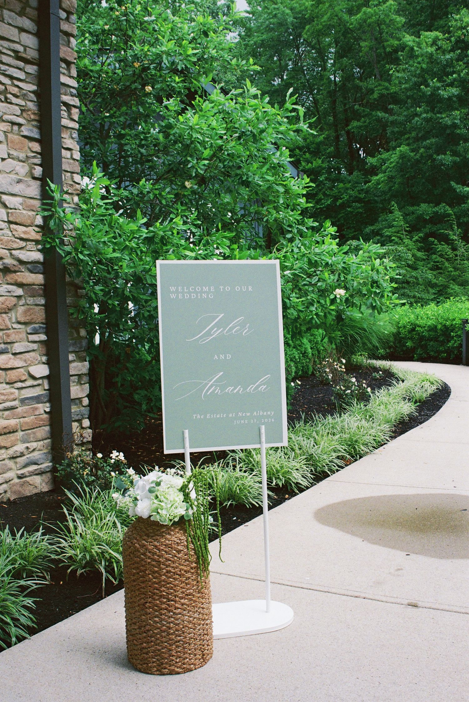 Sage green welcome wedding sign for Tyler and Amanda displayed outdoors with white floral arrangement.