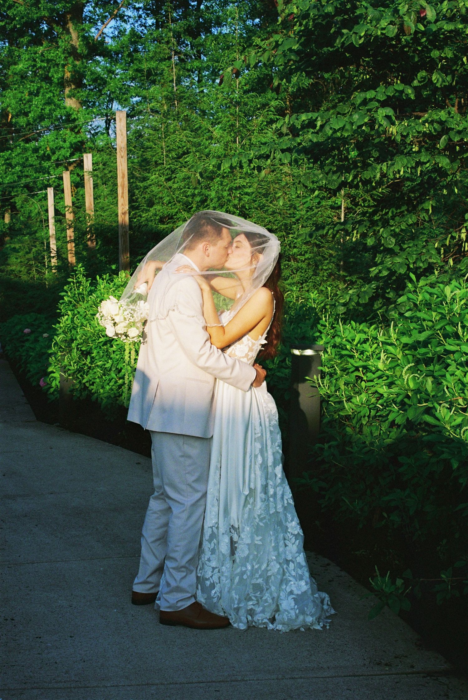 Newlyweds kissing under a bridal veil along a garden pathway at dusk.