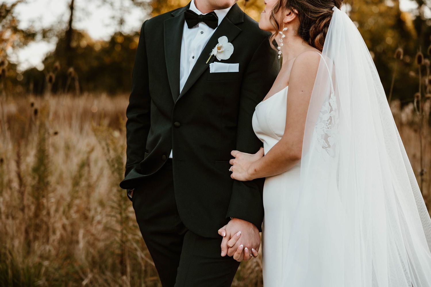 A romantic wedding portrait of a couple in formal attire standing in a golden field at sunset.