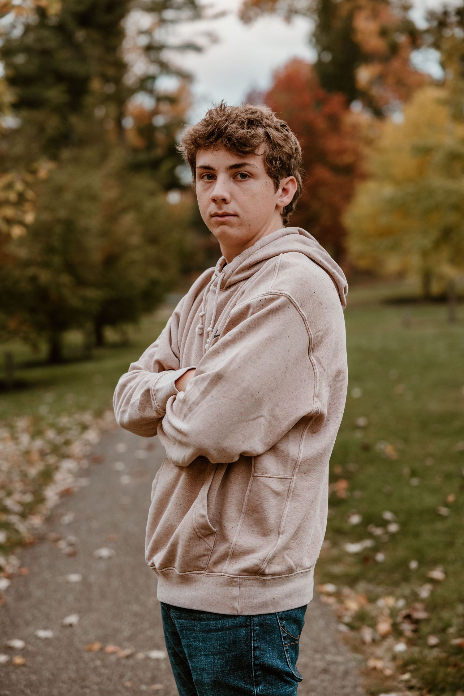 Person in beige hoodie poses outdoors during fall with autumn leaves on ground.