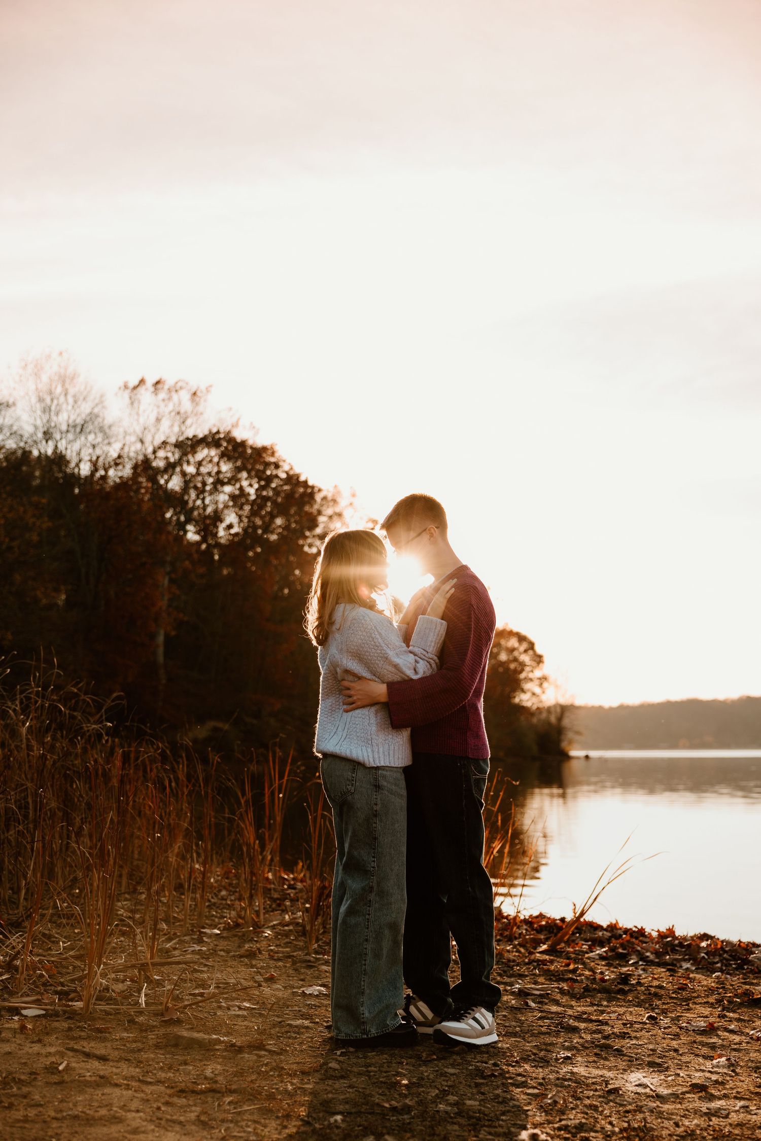 A couple shares an intimate moment at sunset by a lake with golden light streaming through.