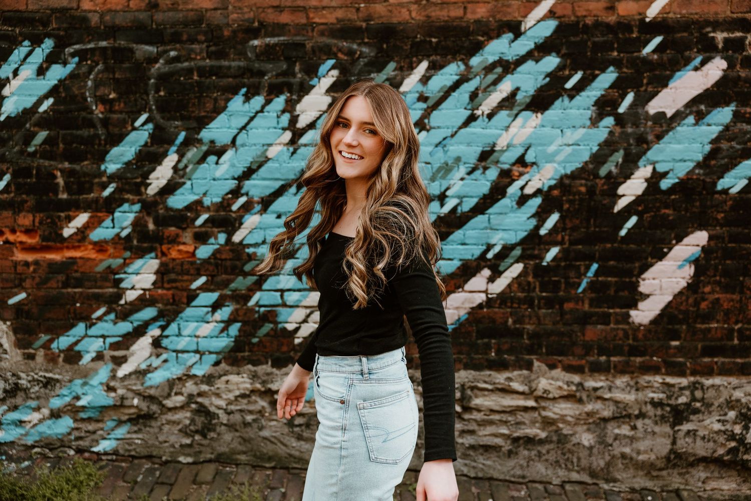 Graduate poses against weathered blue and brick wall in casual attire.