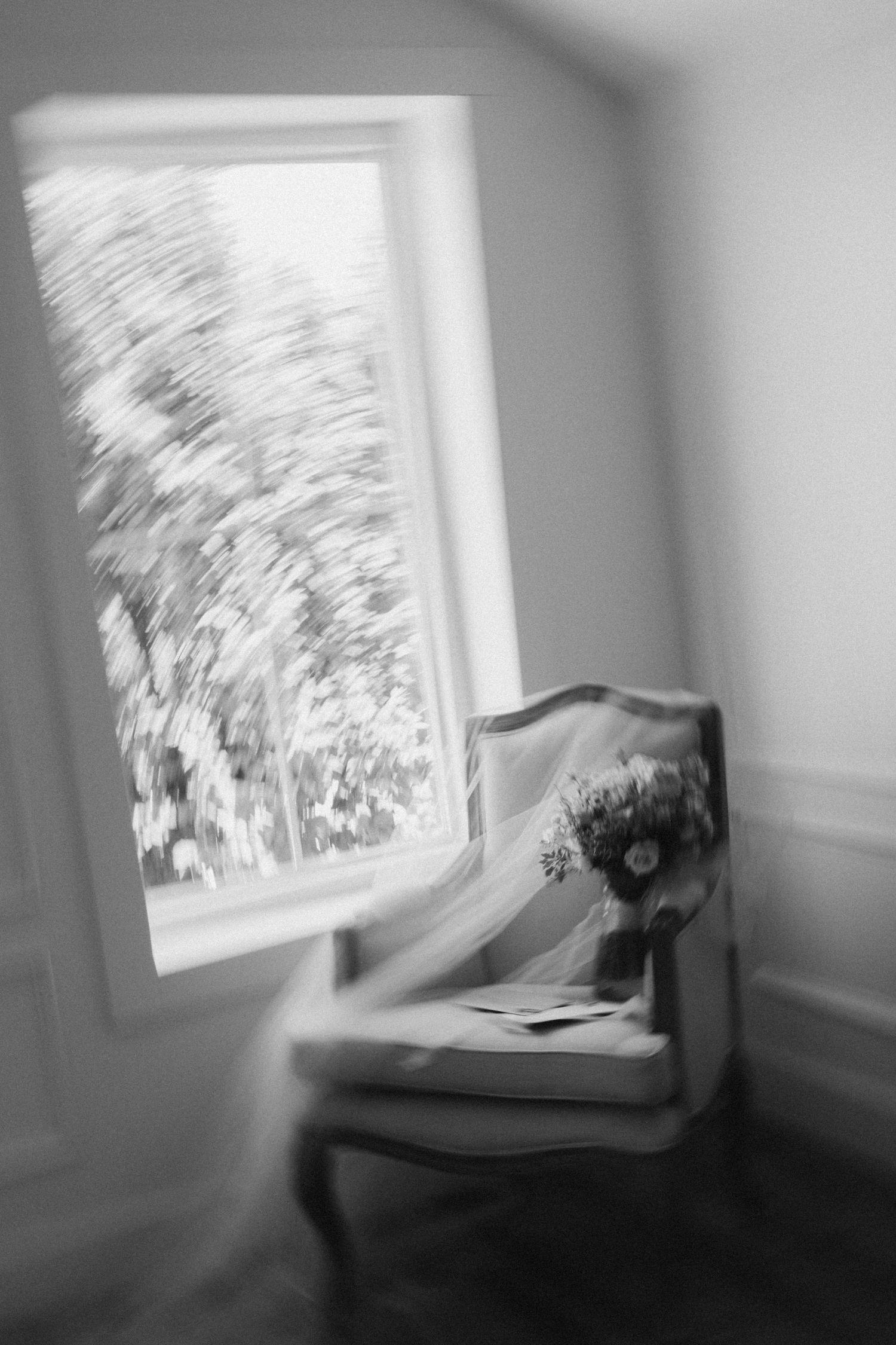 Blurred black and white photo of a vintage chair with a bridal bouquet and veil near a sunlit window.