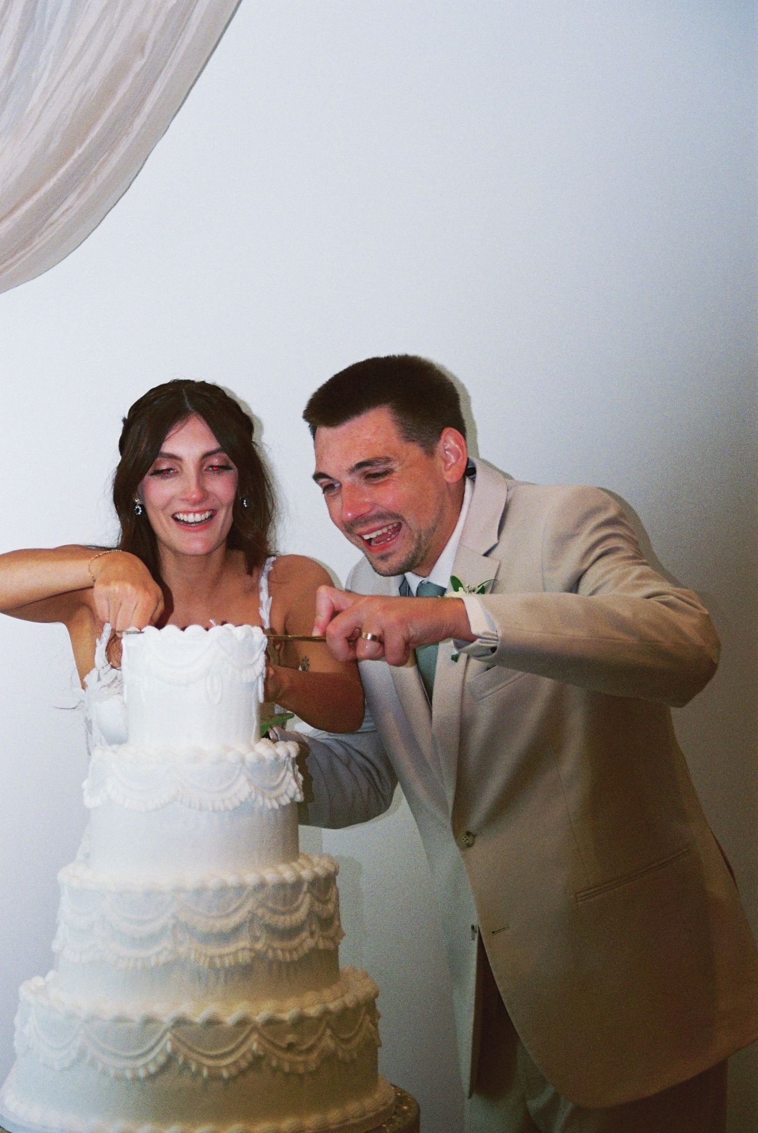 Laughing bride and groom cutting a four-tier white wedding cake together.