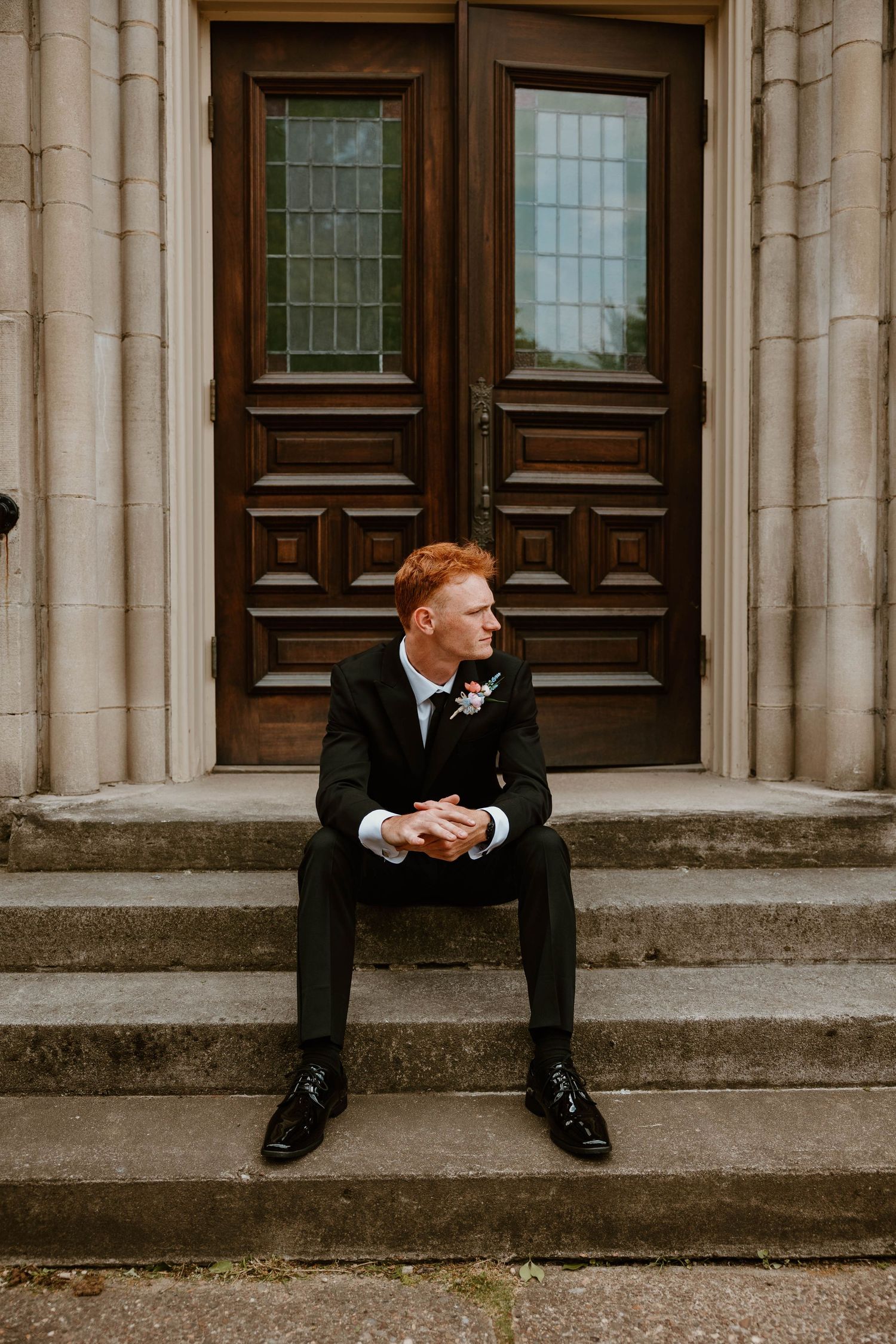 A groom in a dark suit with boutonniere sits on stone steps before ornate wooden doors, looking contemplatively to the side.