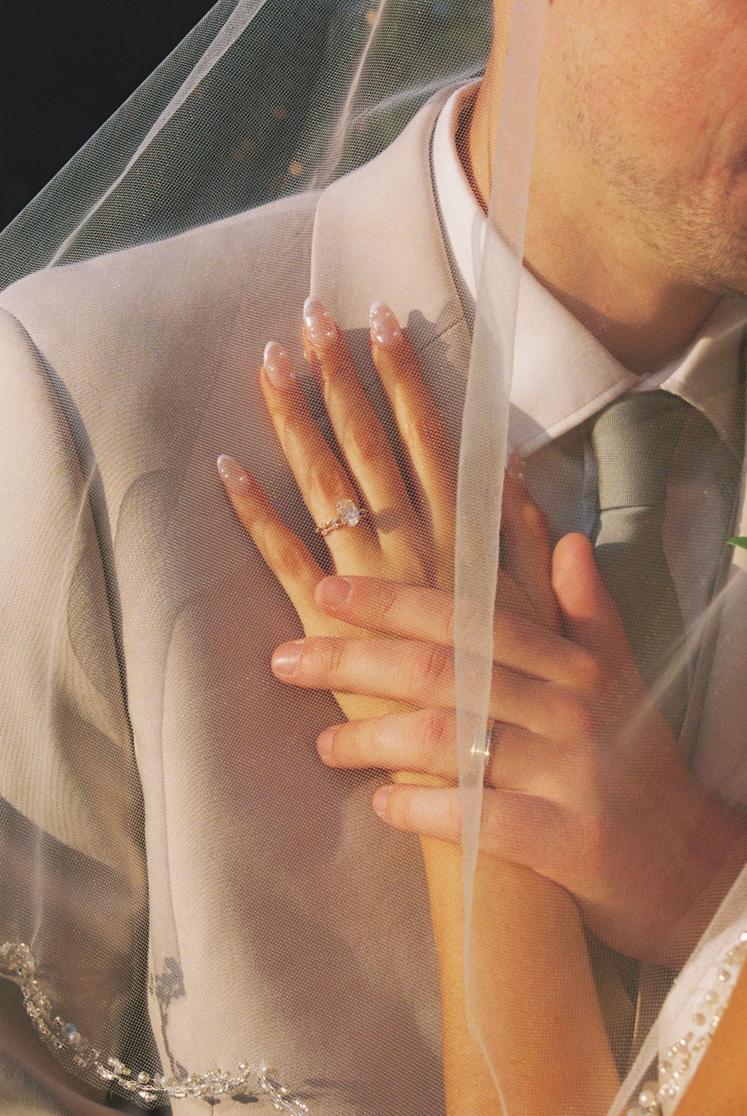 Bride's hand with diamond wedding ring resting on groom's chest in gray suit, partially covered by sheer veil.