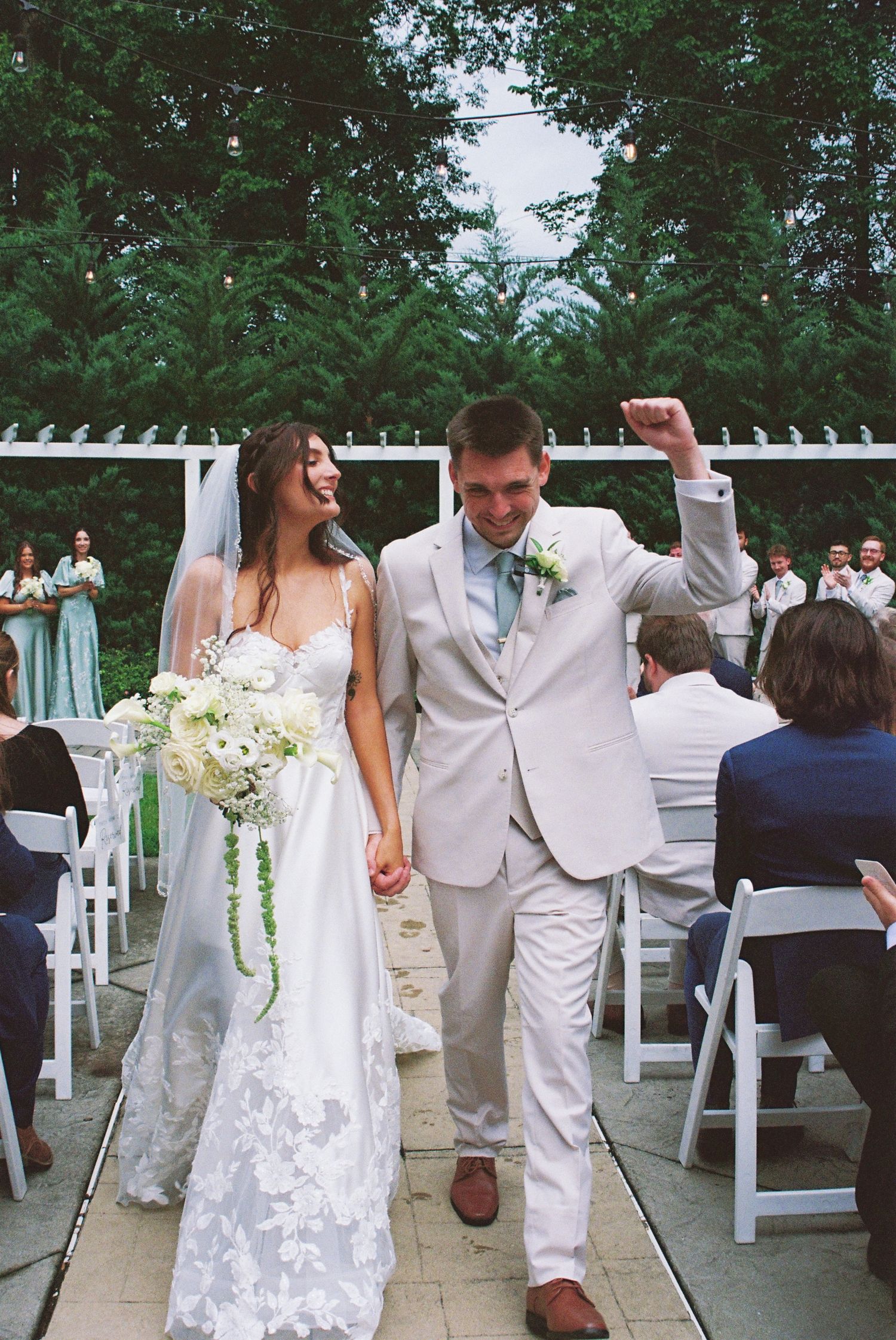 Joyful newlyweds walking down outdoor wedding aisle, groom raising fist in celebration, bride in lace gown.
