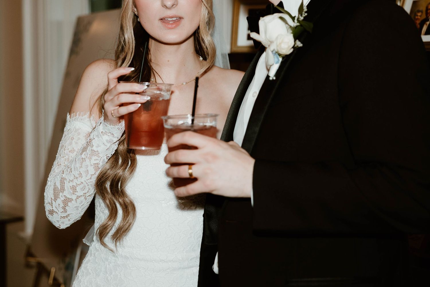 Bride in lace off-shoulder gown and groom in black tuxedo holding cocktails at wedding reception.