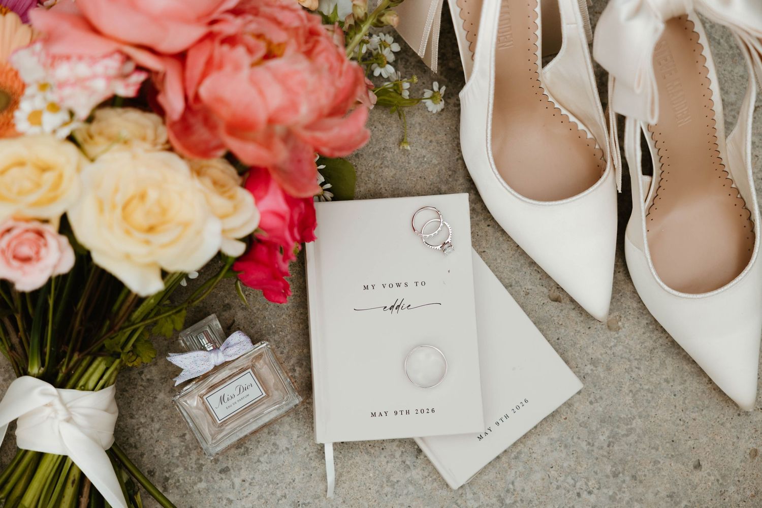 Elegant wedding flat lay with white heels, bridal journal, engagement ring, perfume bottle, and colorful floral bouquet.