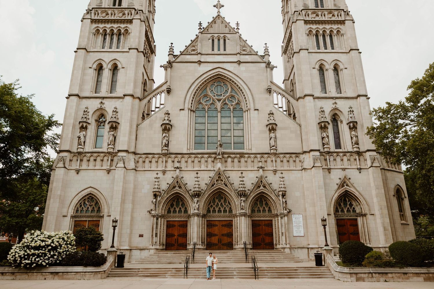 Couple standing before a grand Gothic cathedral with ornate stone arches, large stained glass window, and twin towers.