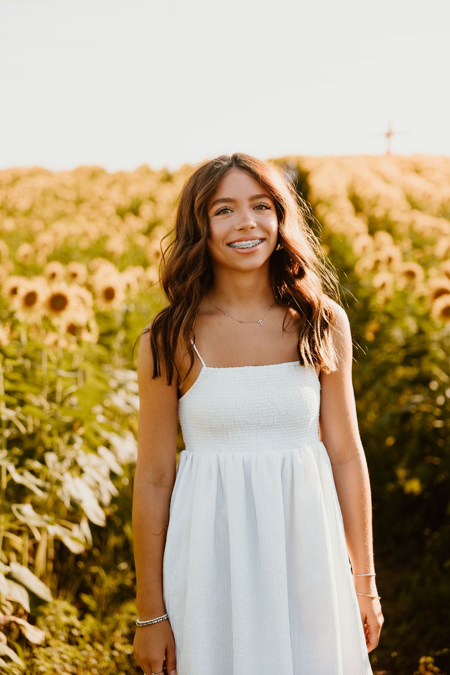 A person wearing a flowing white sundress poses in a sunflower field at sunset.