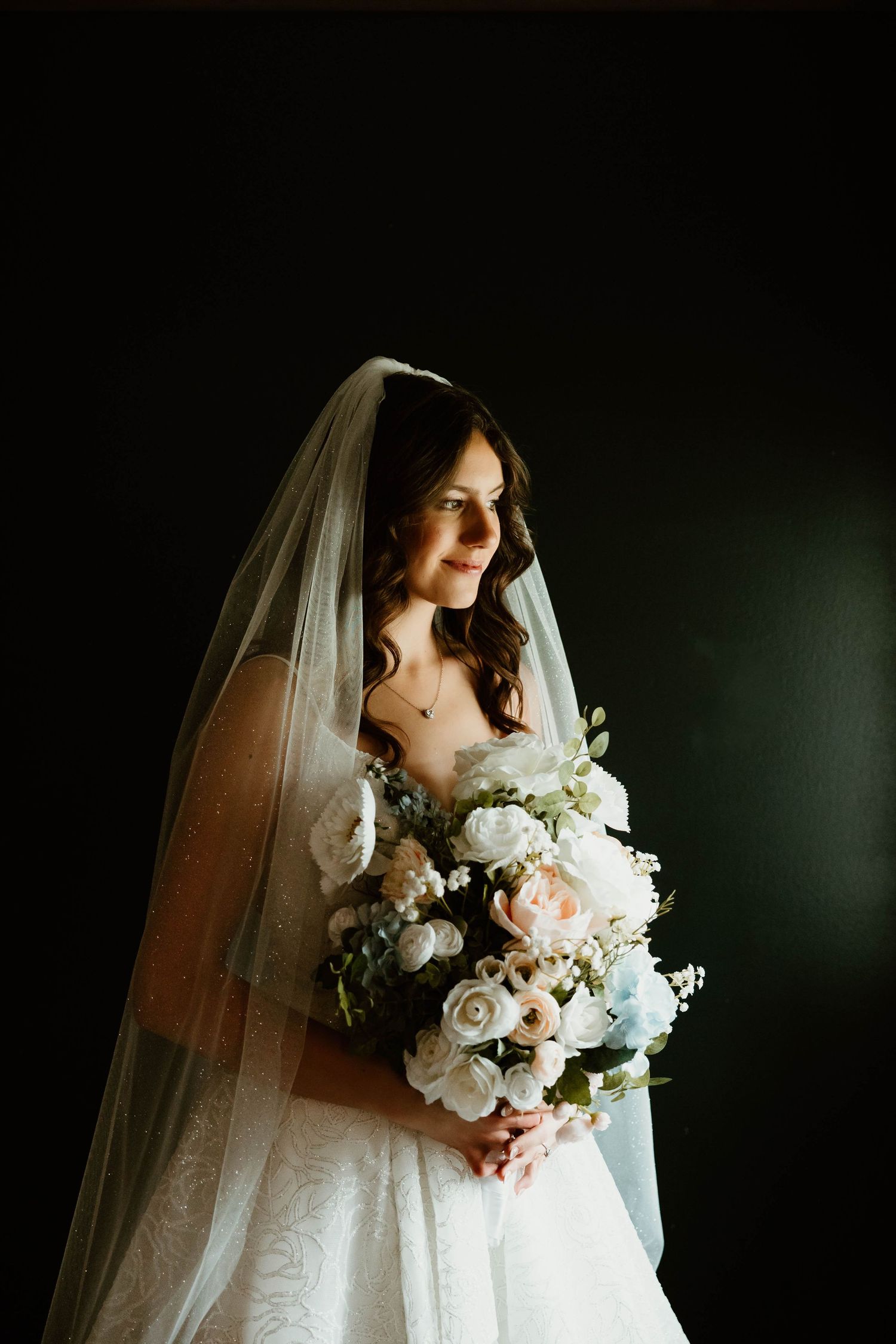 Bride in white lace gown and veil holds elegant bouquet of white and blush roses against dark background.