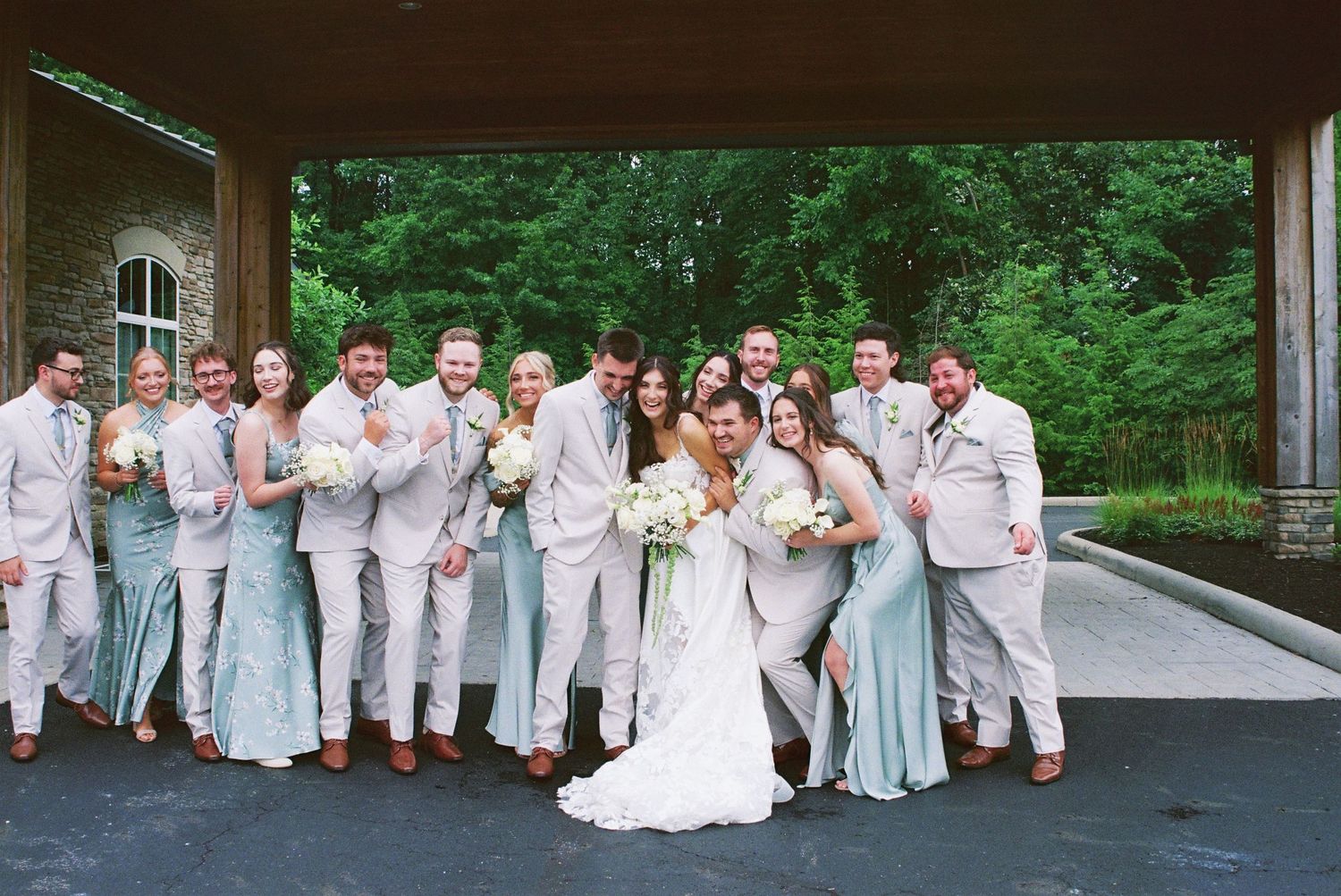 Wedding party in sage green bridesmaid dresses and light gray suits posing under a stone pavilion.