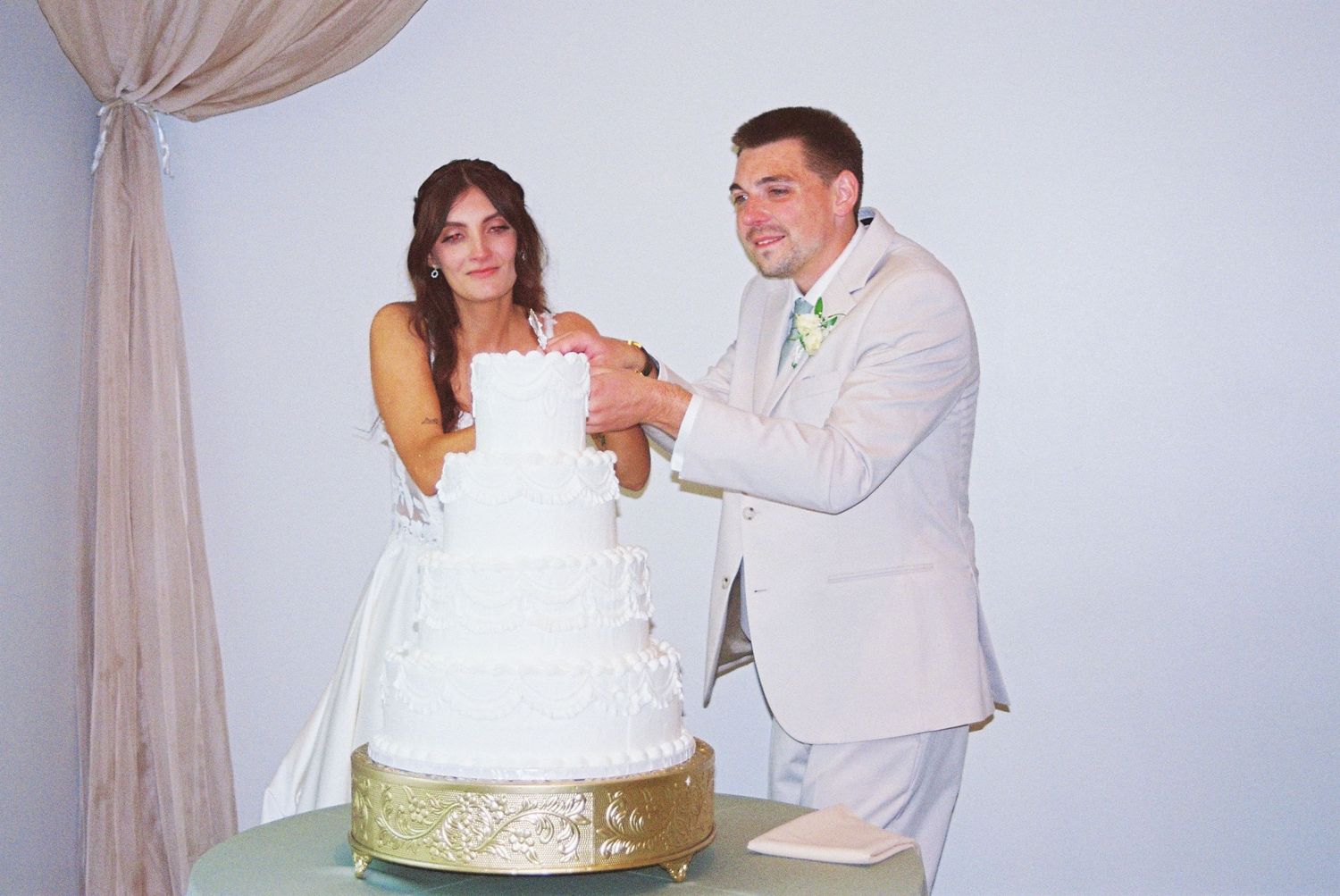 Smiling bride and groom cut elegant four-tier white wedding cake with gold decorative stand.