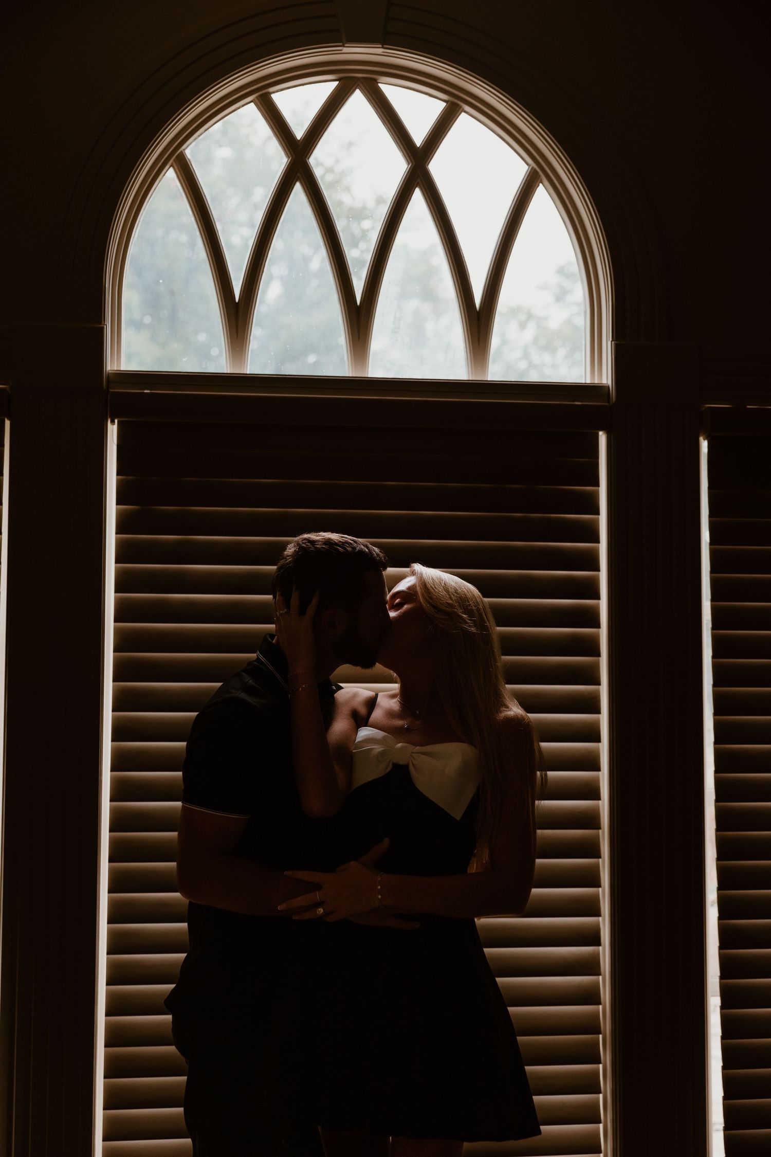 A silhouetted couple embraces near an arched window with venetian blinds.