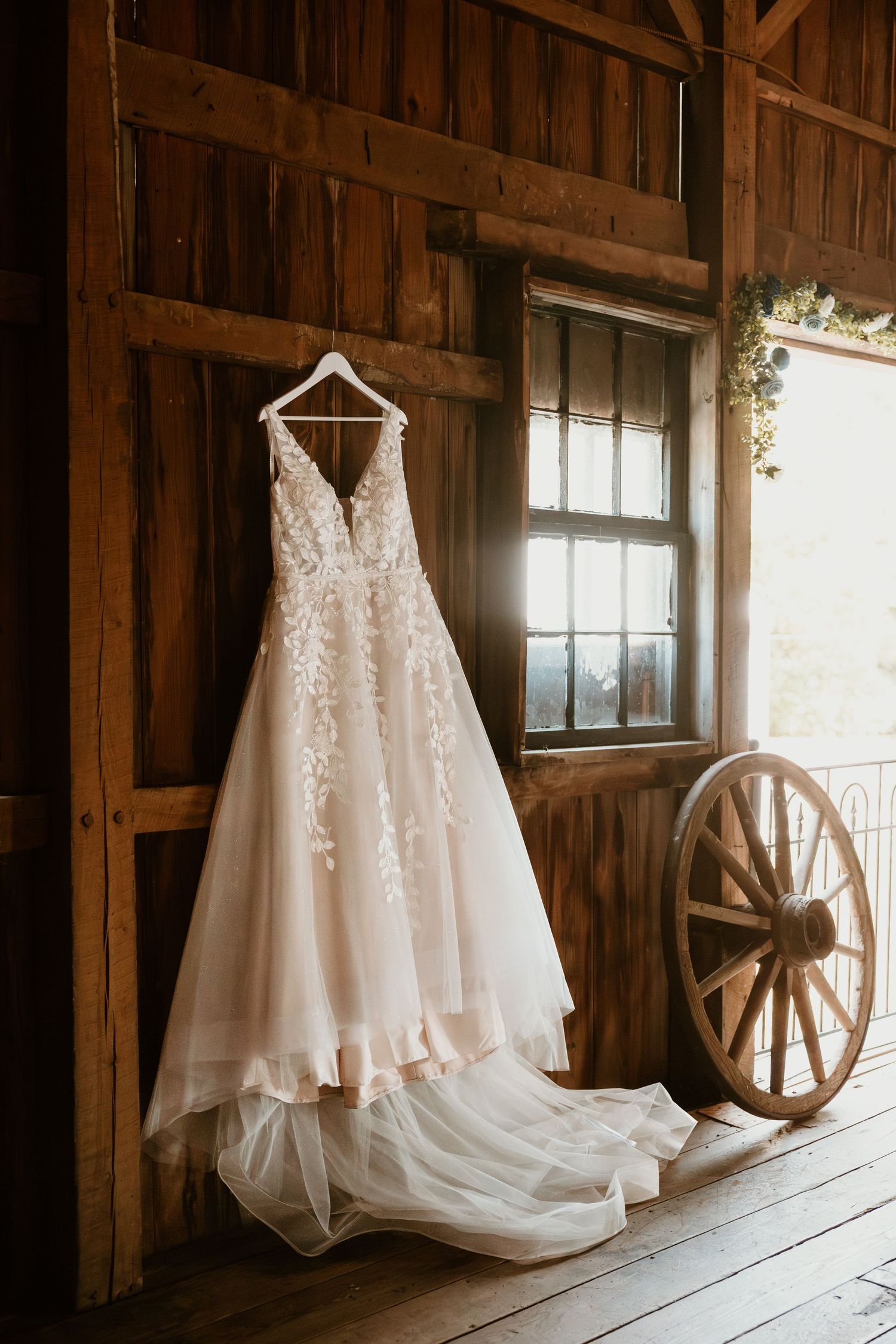 Elegant white lace wedding dress hangs in rustic barn setting with vintage wagon wheel decor.