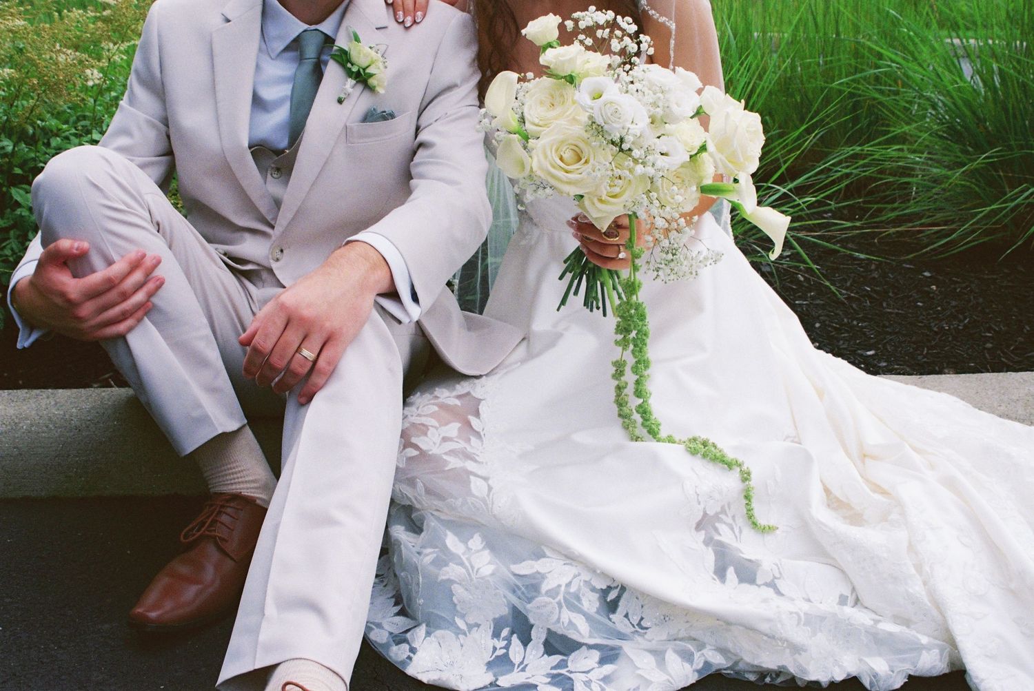 Bride in lace gown and groom in tan suit sitting together holding a cascading white rose bouquet.
