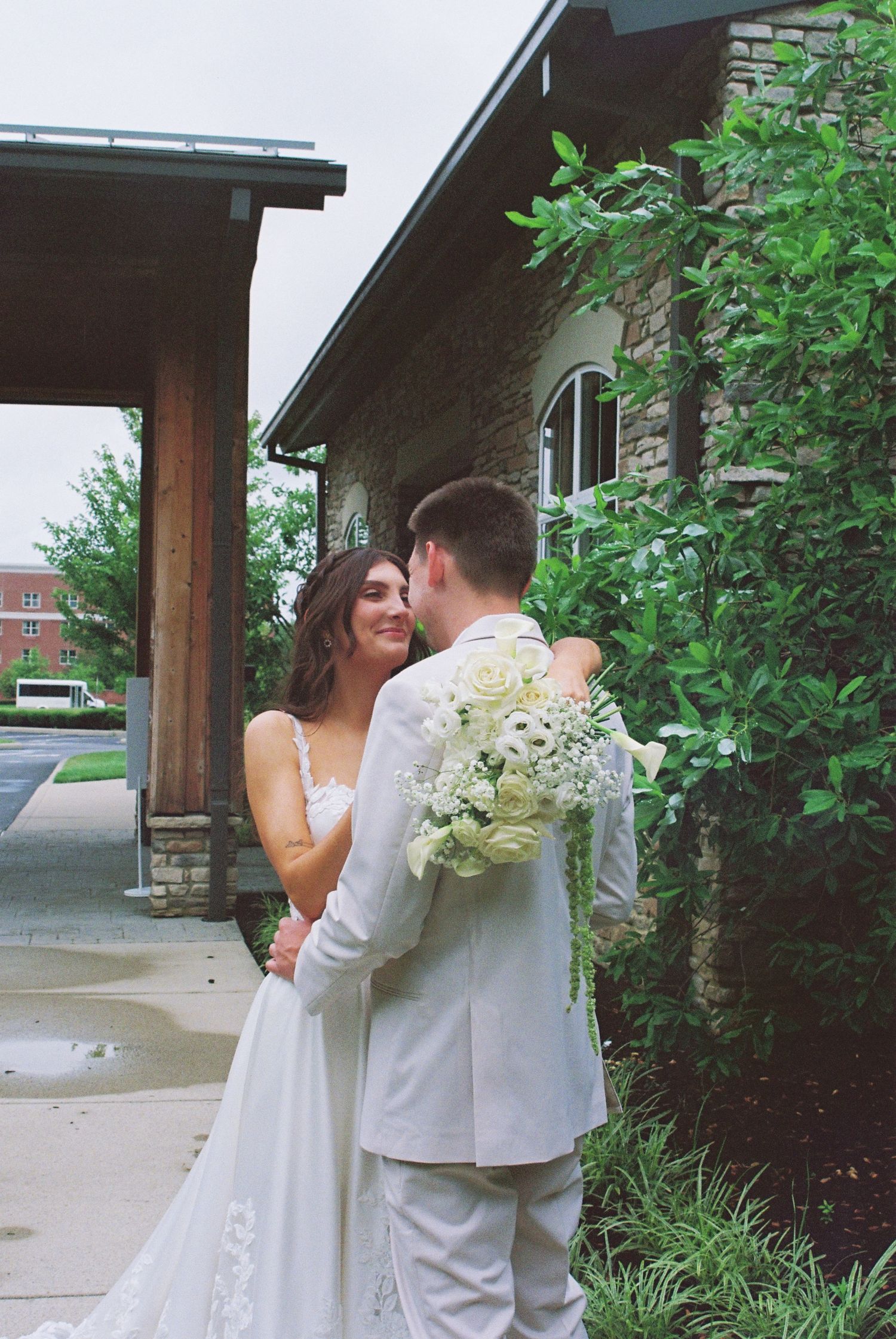 Bride and groom share a romantic embrace outside a stone building, bride holding white floral bouquet.