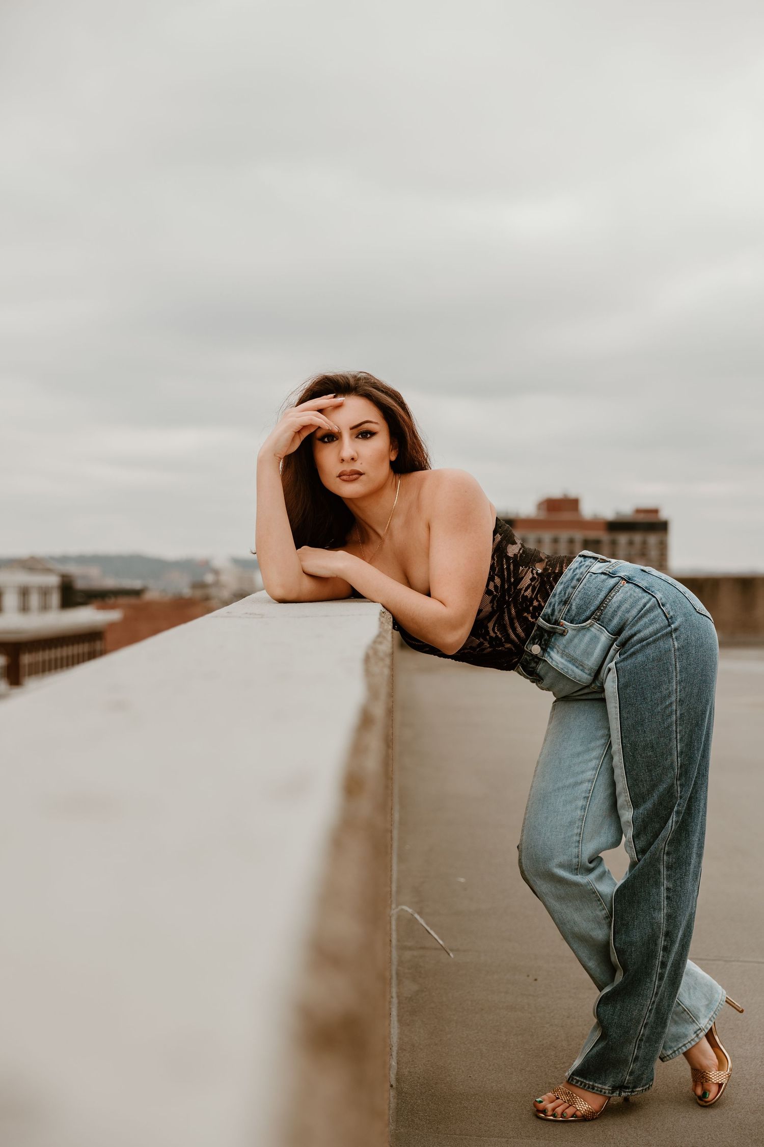 Woman in casual denim leans against a rooftop wall with cityscape in background.
