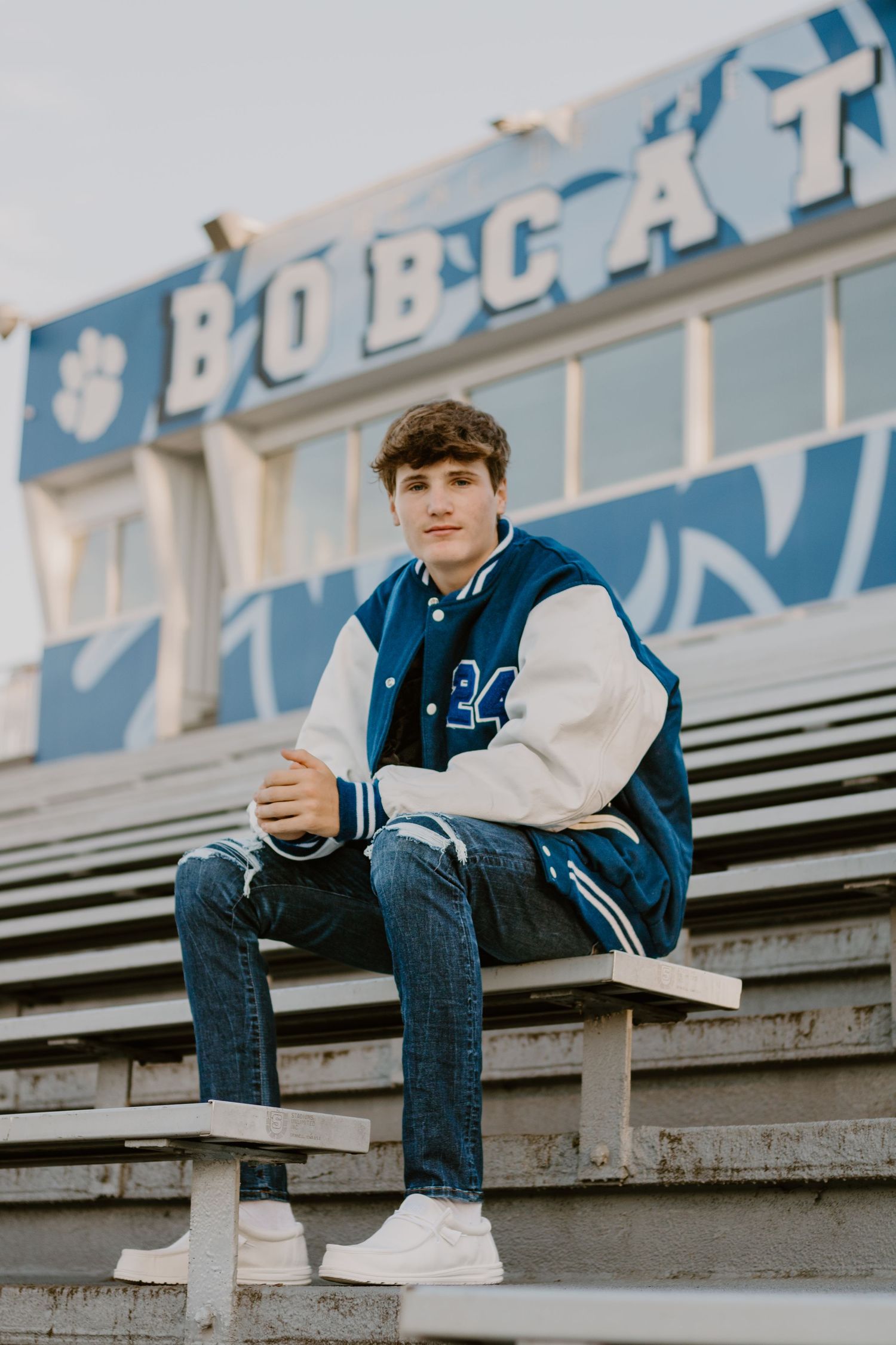 Student sitting on bleachers wearing varsity jacket and jeans in casual outdoor photo shoot.