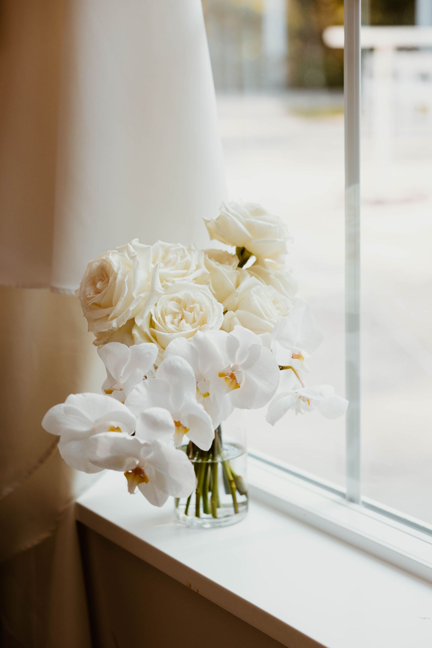 White orchids and roses arranged in clear vase sit elegantly on windowsill.