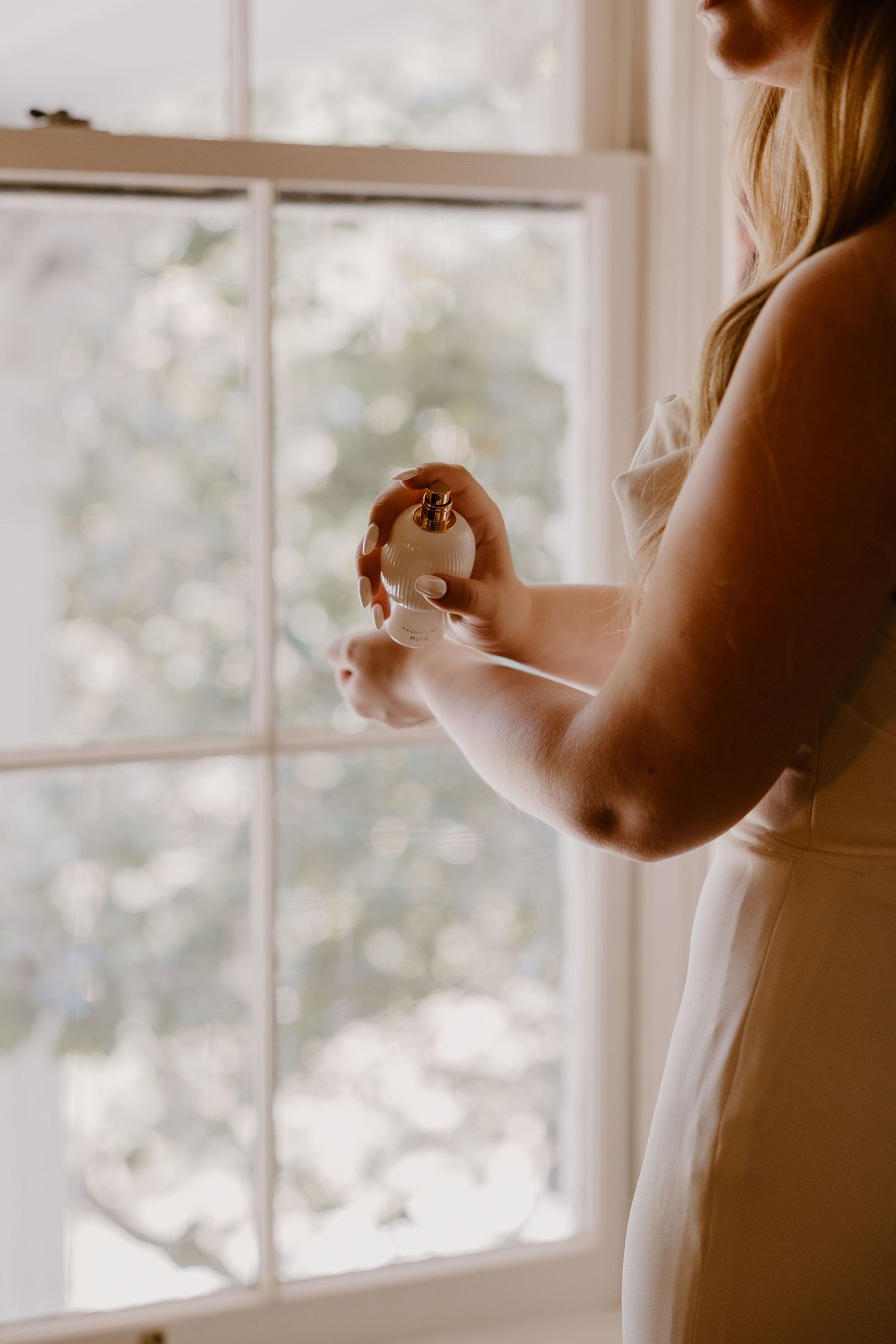 Bride in white dress adjusting jewelry by sunlit window.