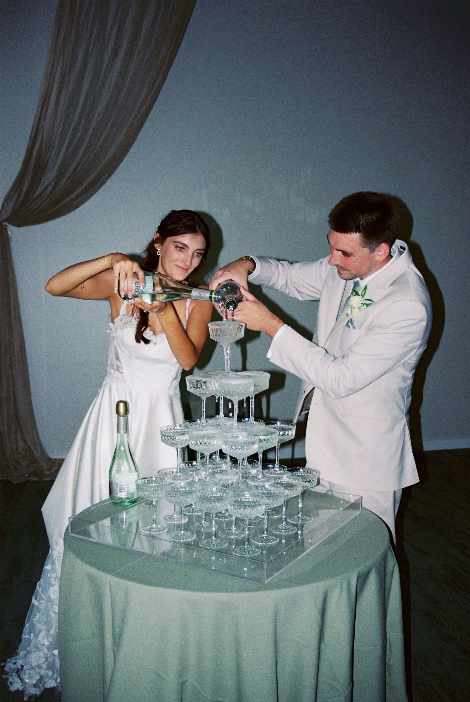 Bride and groom pour champagne together into elegant crystal tower of coupe glasses at reception.