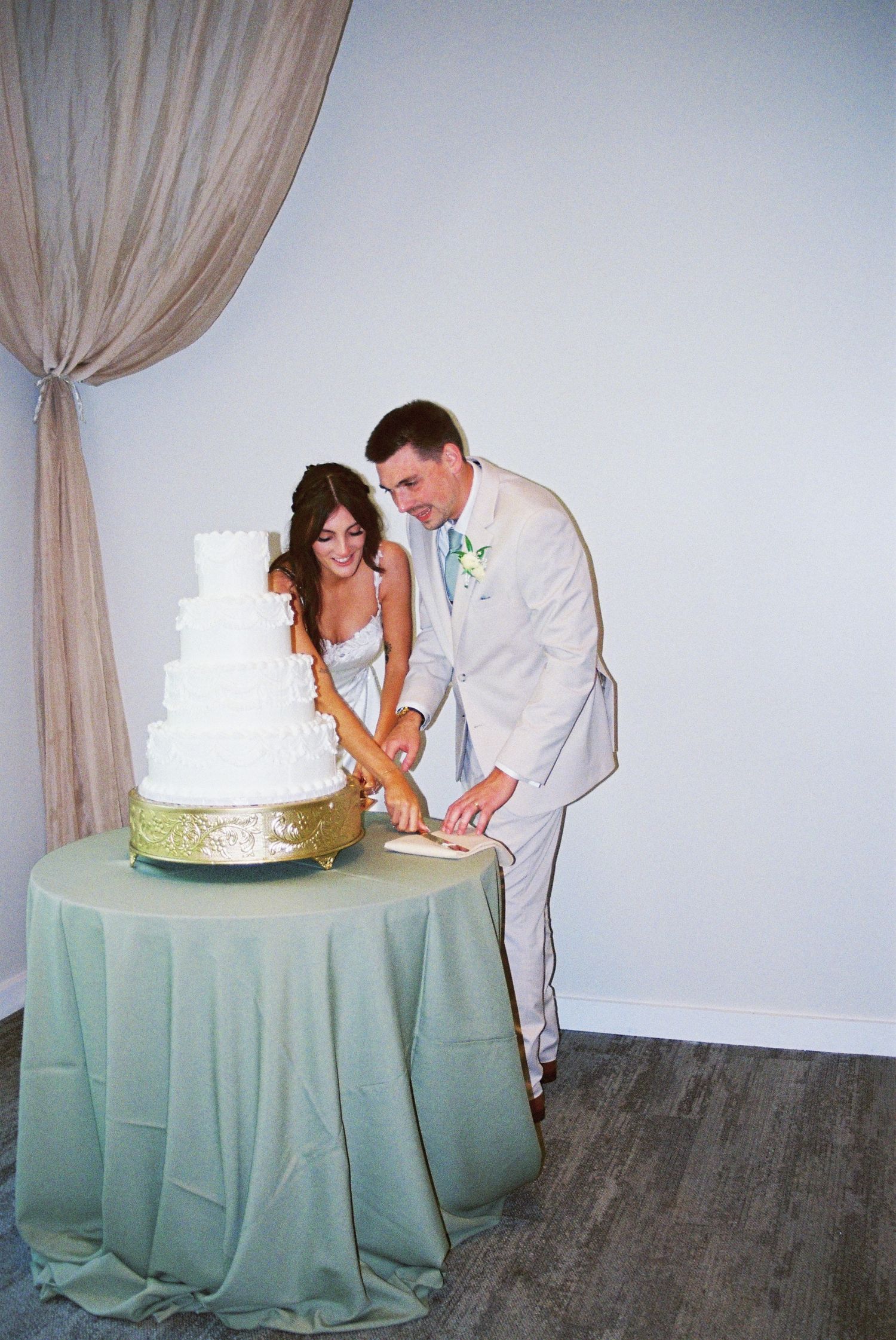Bride and groom in ivory suit cutting a four-tier white wedding cake on a sage green tablecloth.