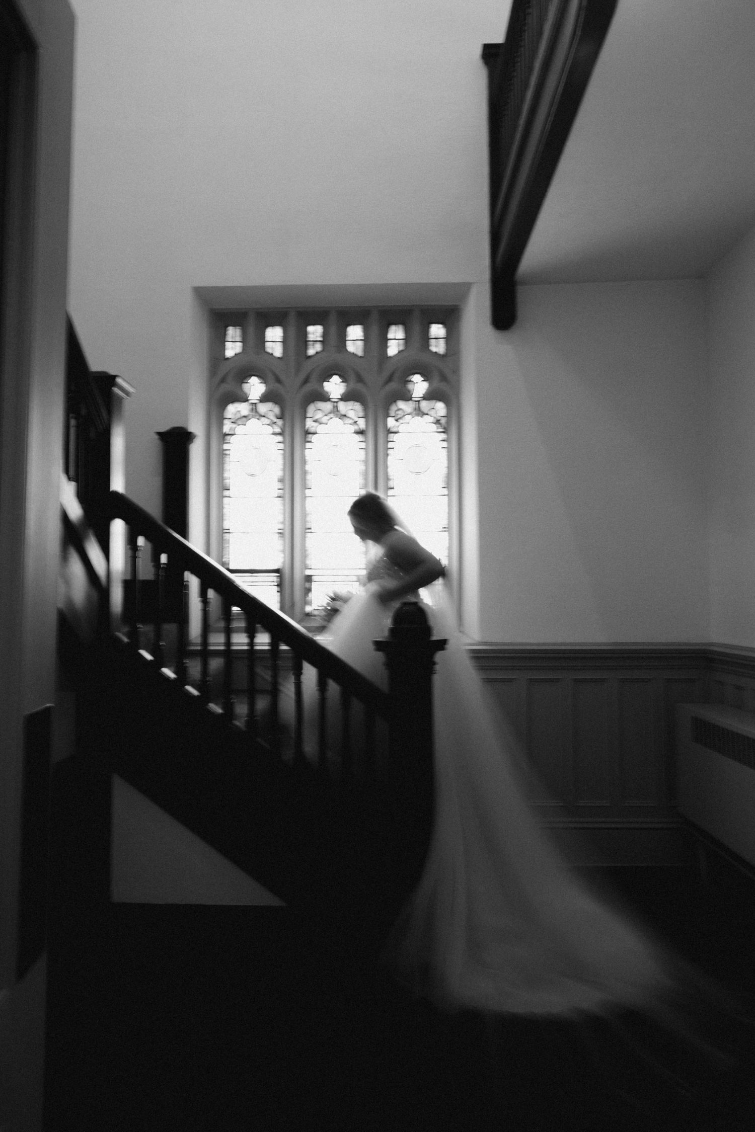 Black and white photo of a bride in a flowing gown descending an elegant staircase near a Gothic arched window.