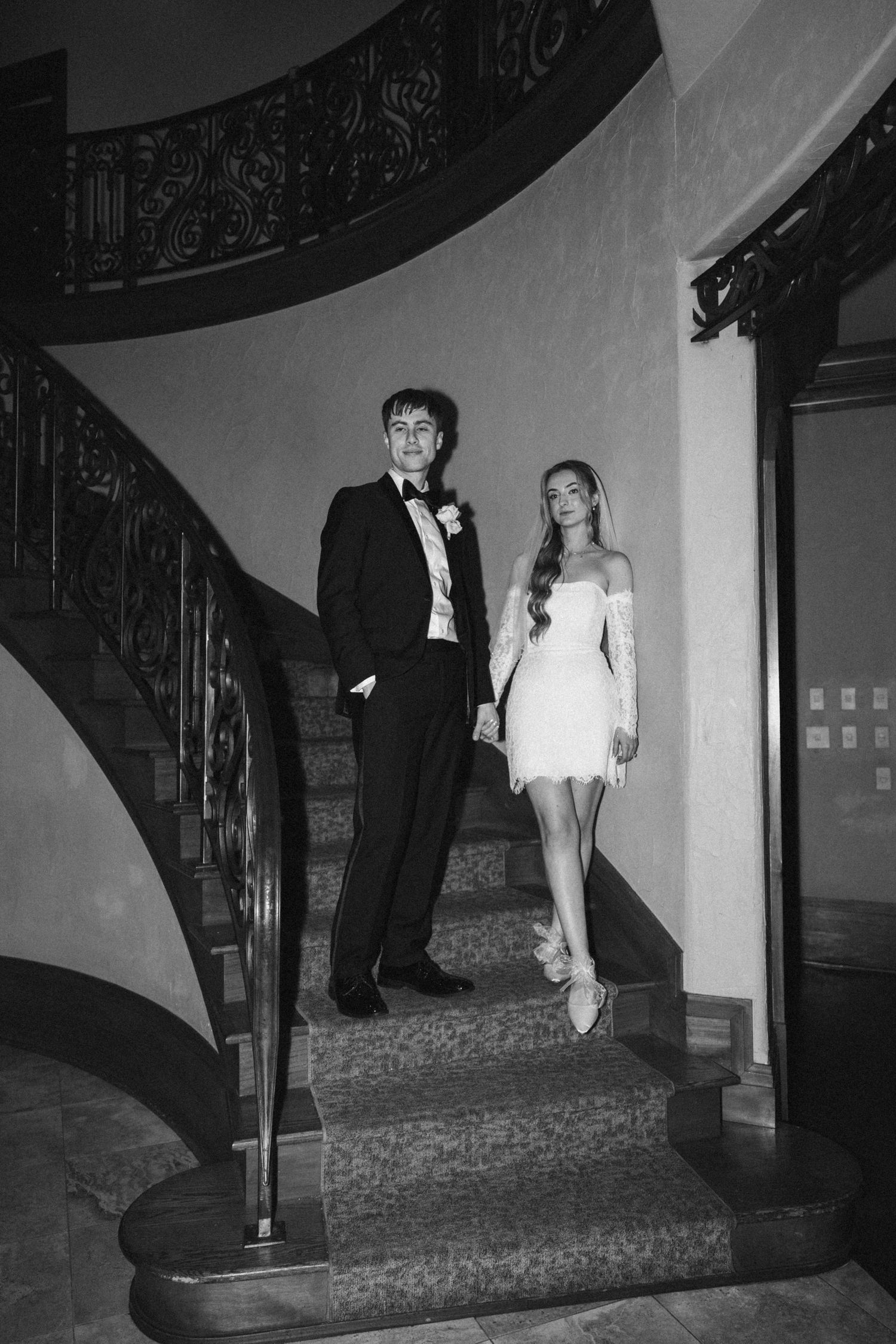 Black and white photo of elegant couple in wedding attire posing on grand spiral staircase.