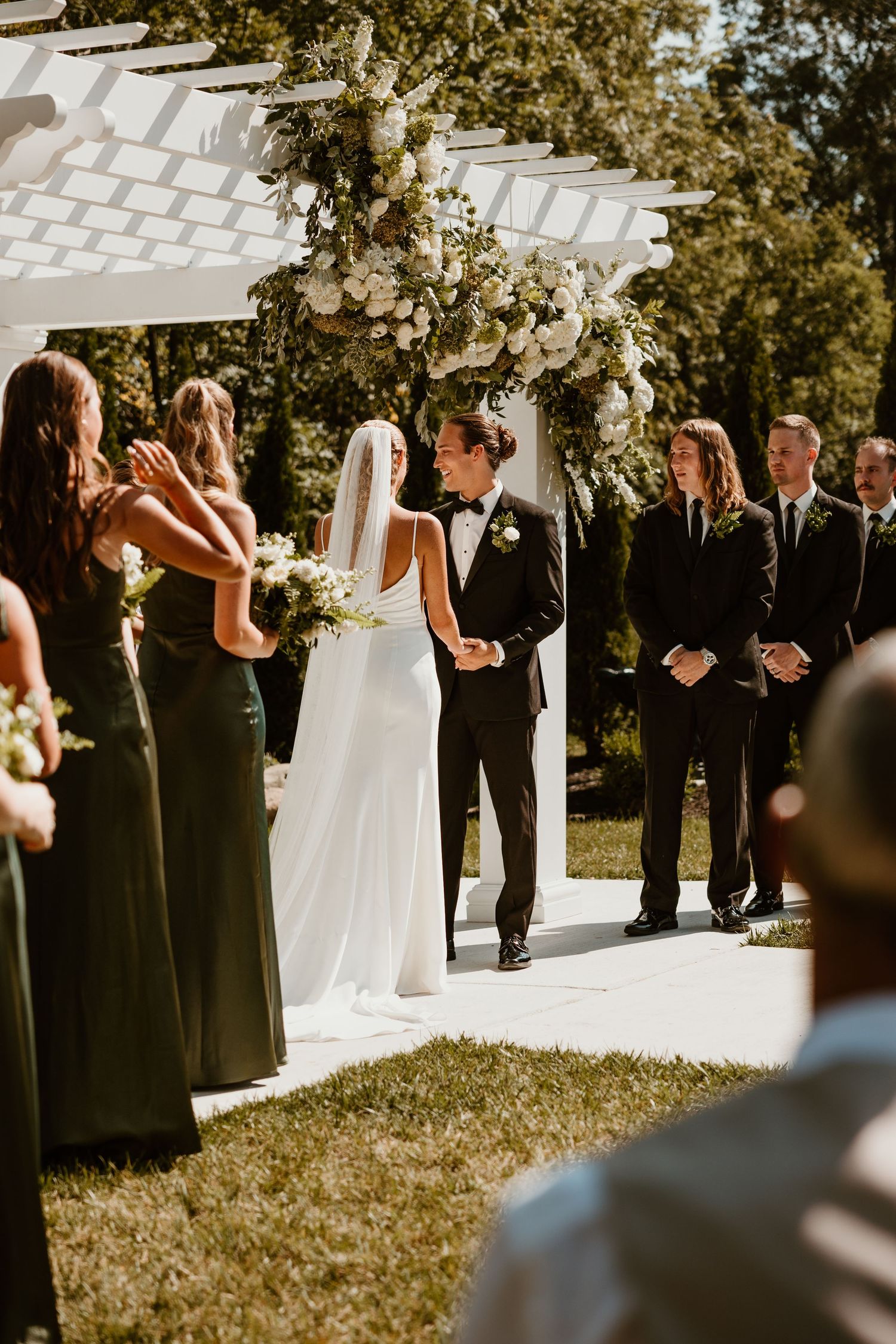 Wedding party in black attire stands outdoors in front of white pergola during sunny ceremony.