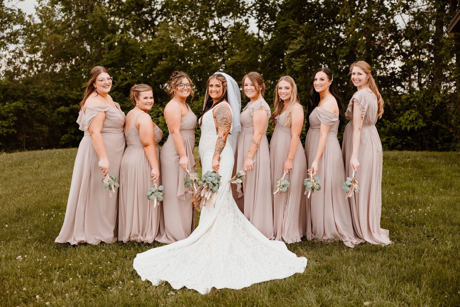 Bridesmaids in soft pink dresses pose together on a grassy hill during an outdoor wedding celebration.