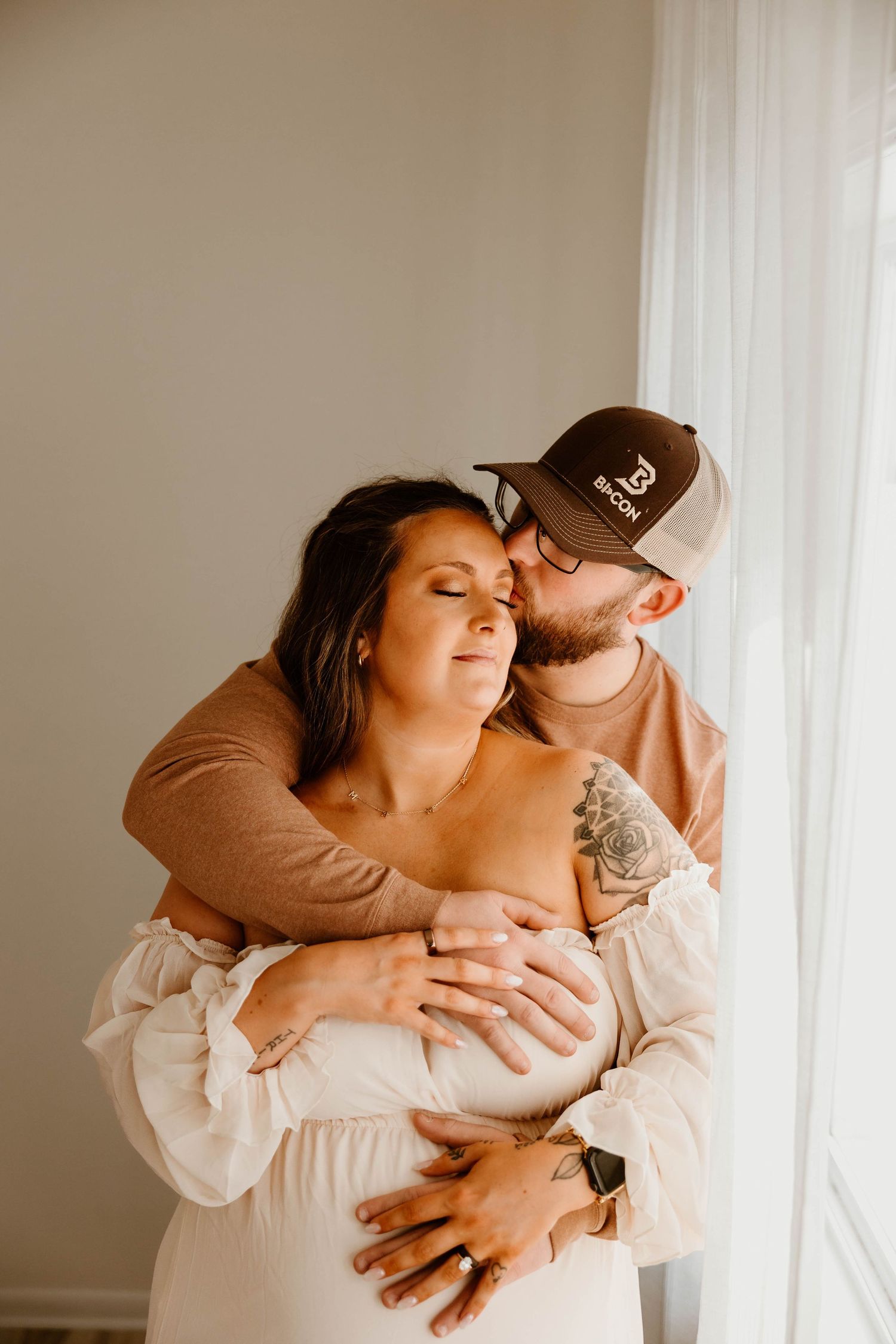 Intimate couple embracing in sunlit room with white curtains.