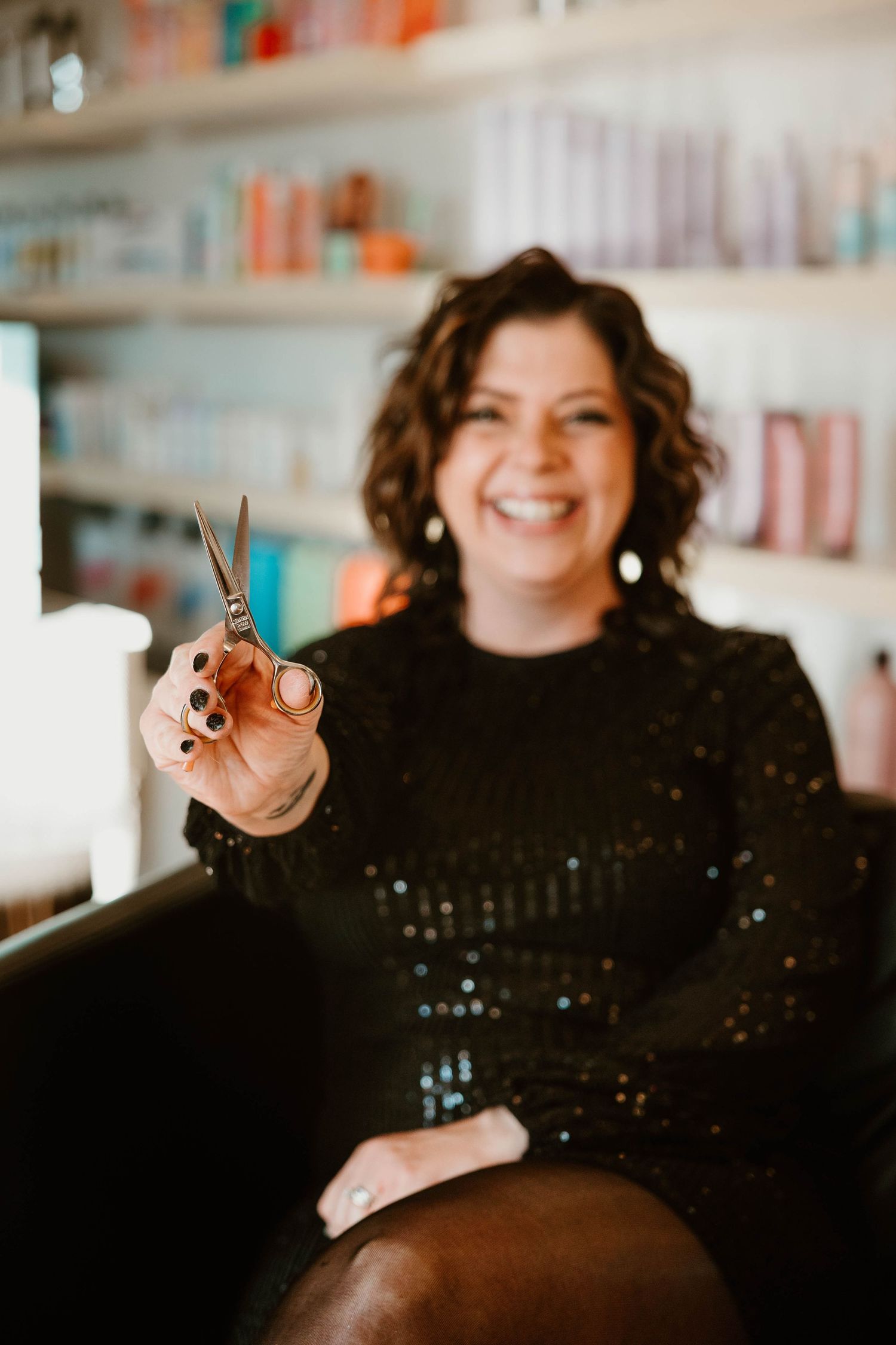 Salon professional in black sequin top holding styling tools against blurred product shelves.