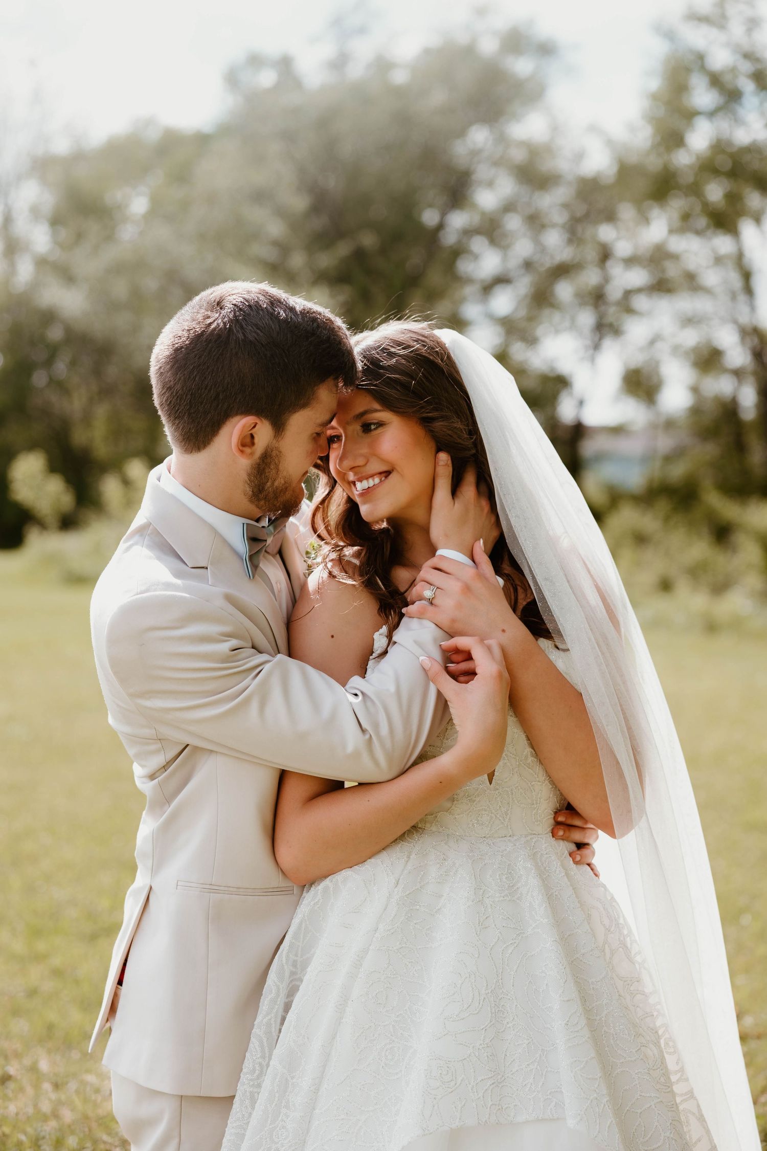 A smiling bride in a white gown and veil embraces her groom in a beige suit outdoors on their wedding day.