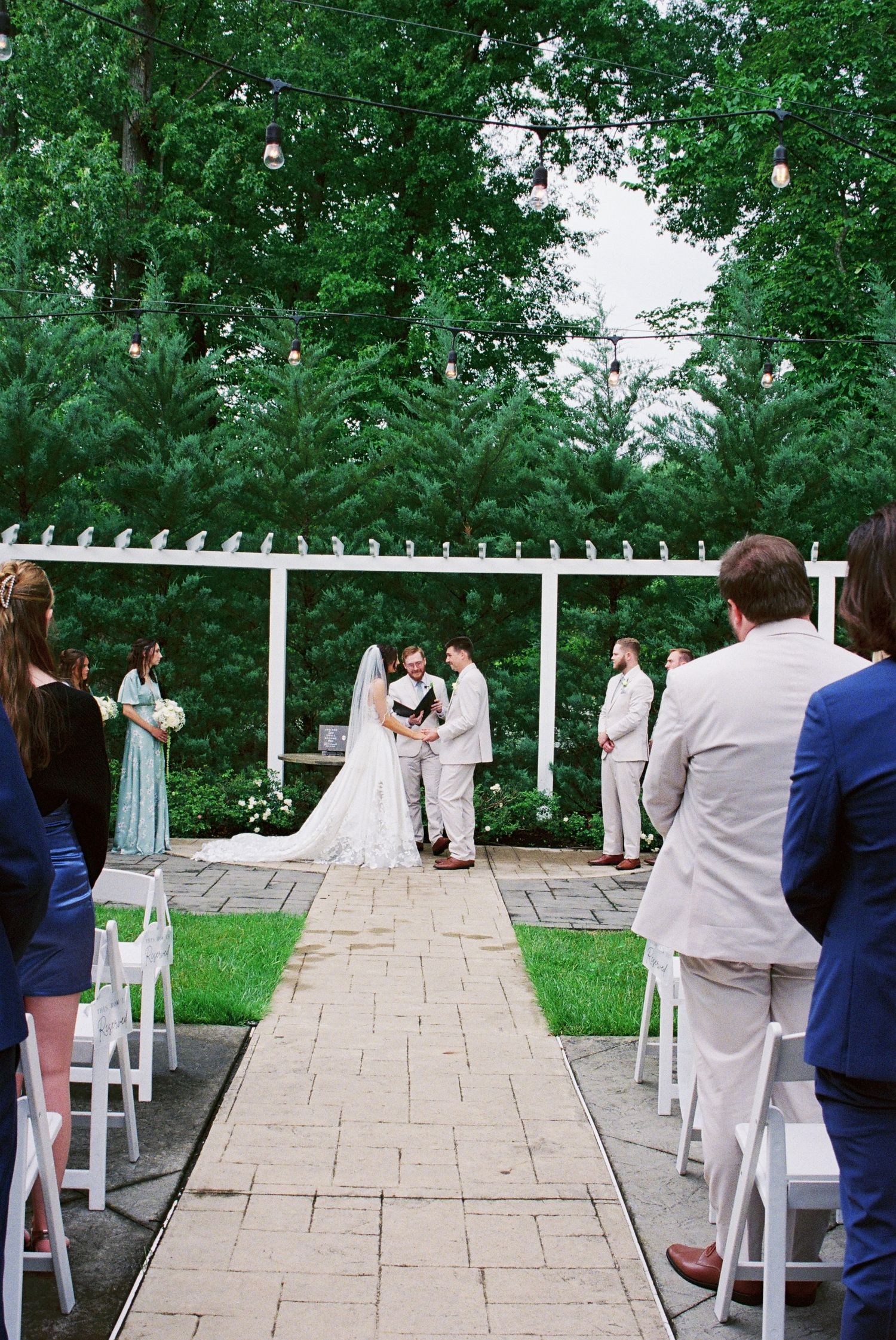 Outdoor wedding ceremony at white pergola altar surrounded by lush green trees.