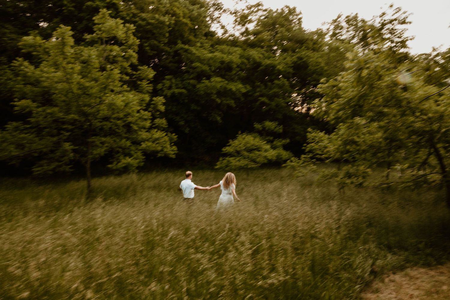 Couple holding hands walking through tall green meadow grass surrounded by lush trees.