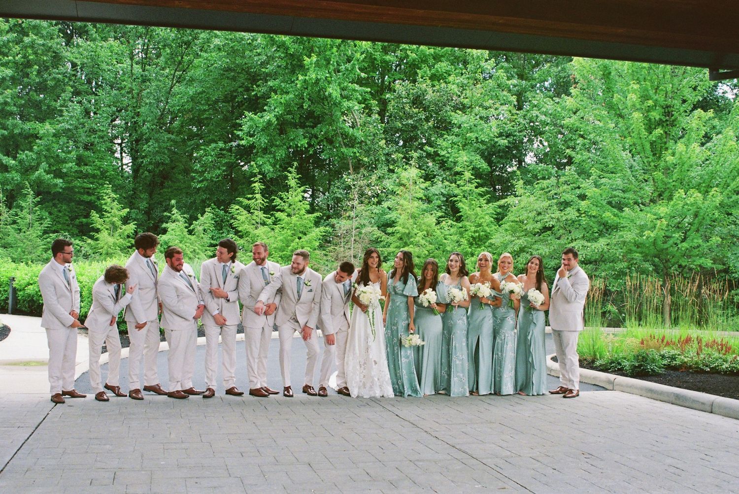Large wedding party in mint bridesmaid gowns and ivory suits lined up outdoors in lush greenery.